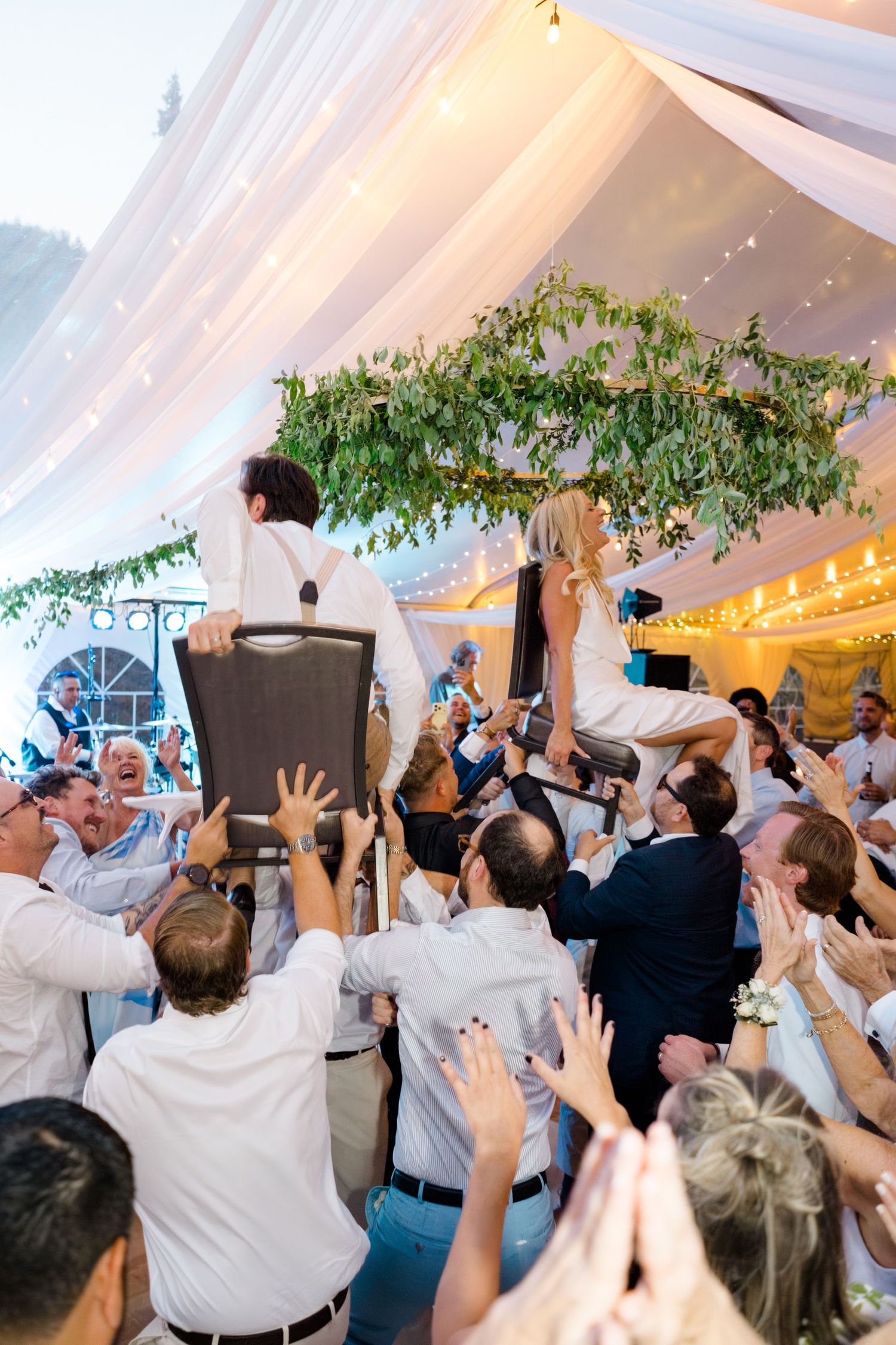 Bride and groom lifted on chairs by guests during lively Jewish Hora dance at elegant tented wedding reception.