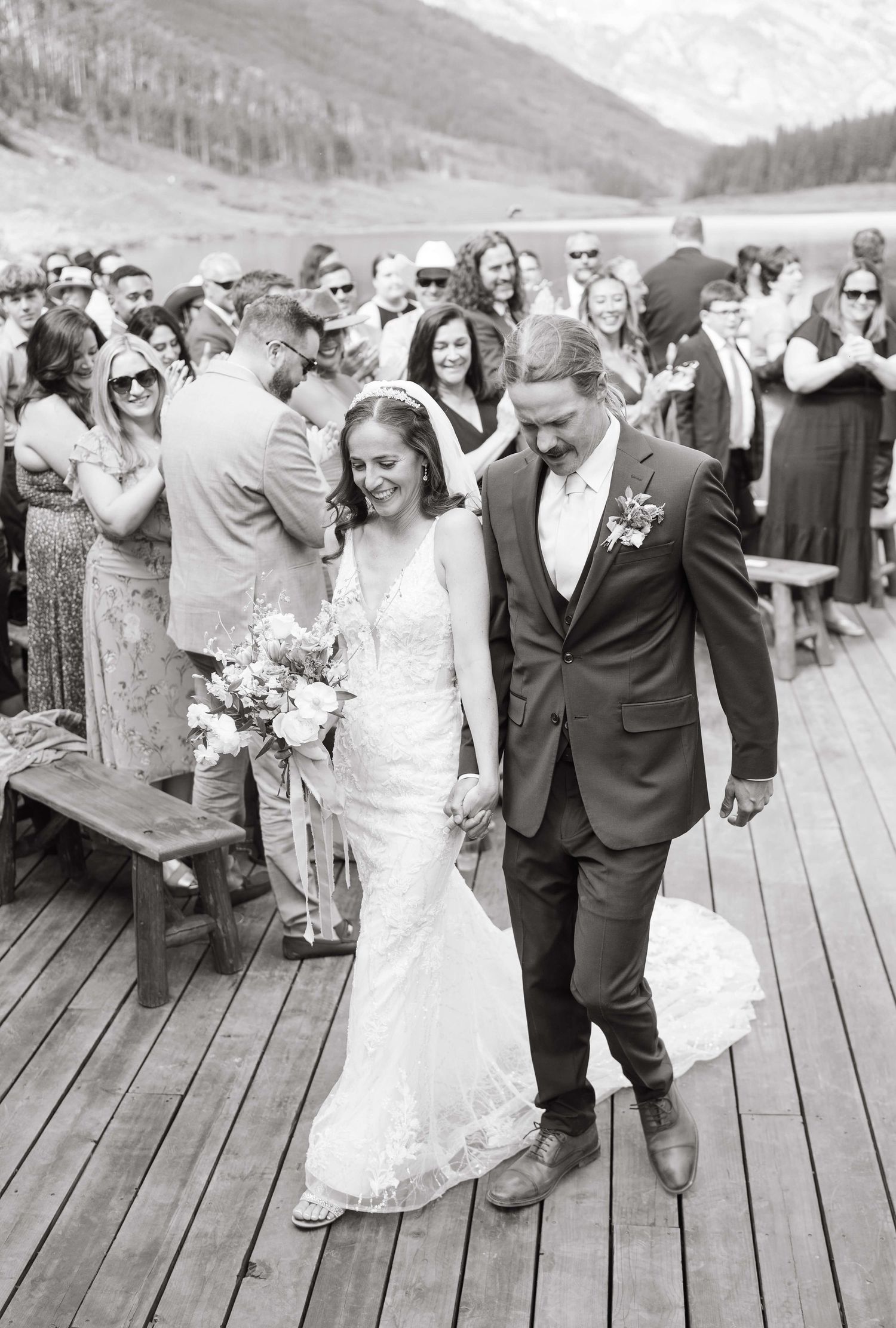 Black and white photo of bride and groom walking down the aisle on a wooden deck with mountain lake backdrop at piney river ranch wedding.