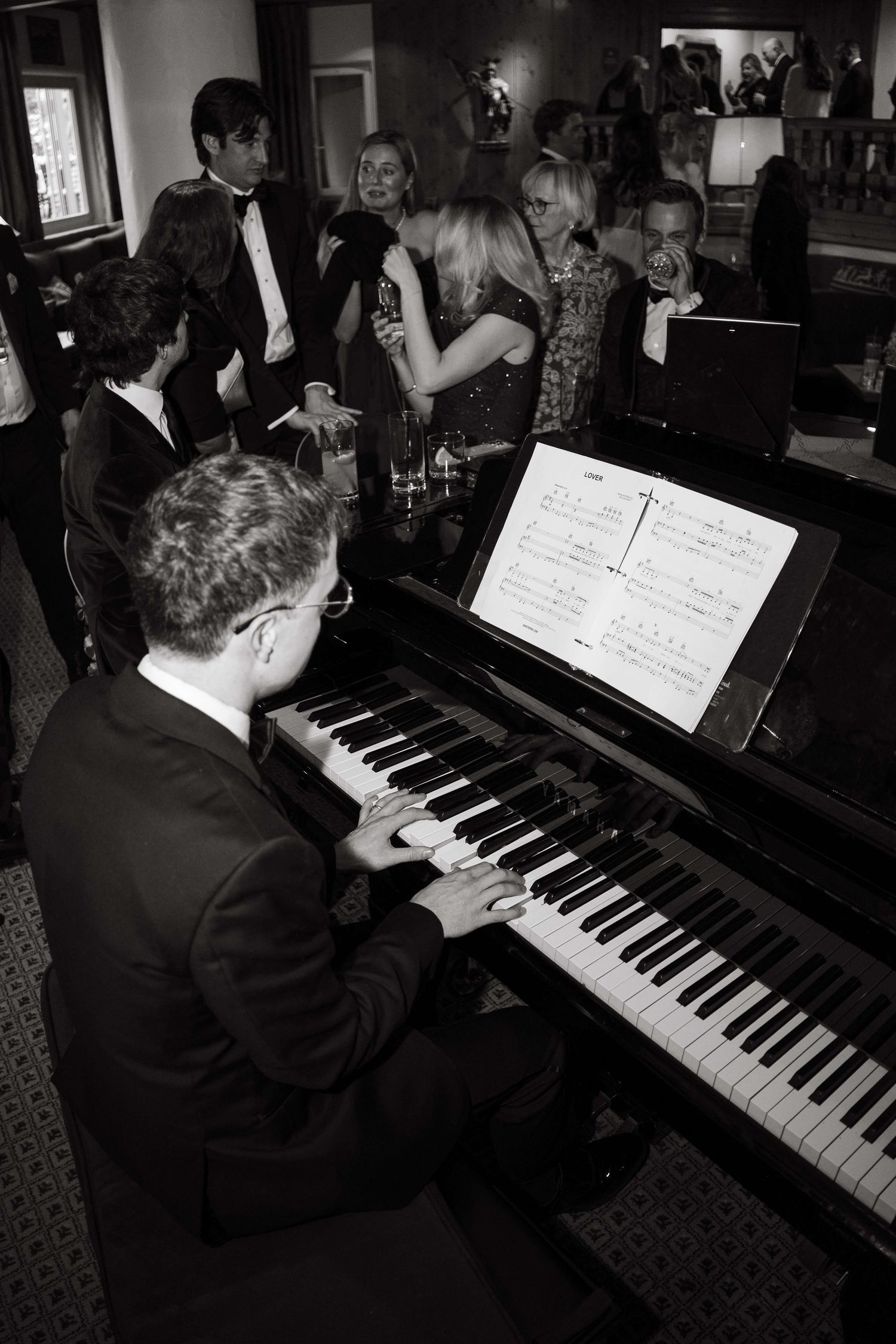 A pianist in a suit plays grand piano at an elegant party while guests socialize in the background.