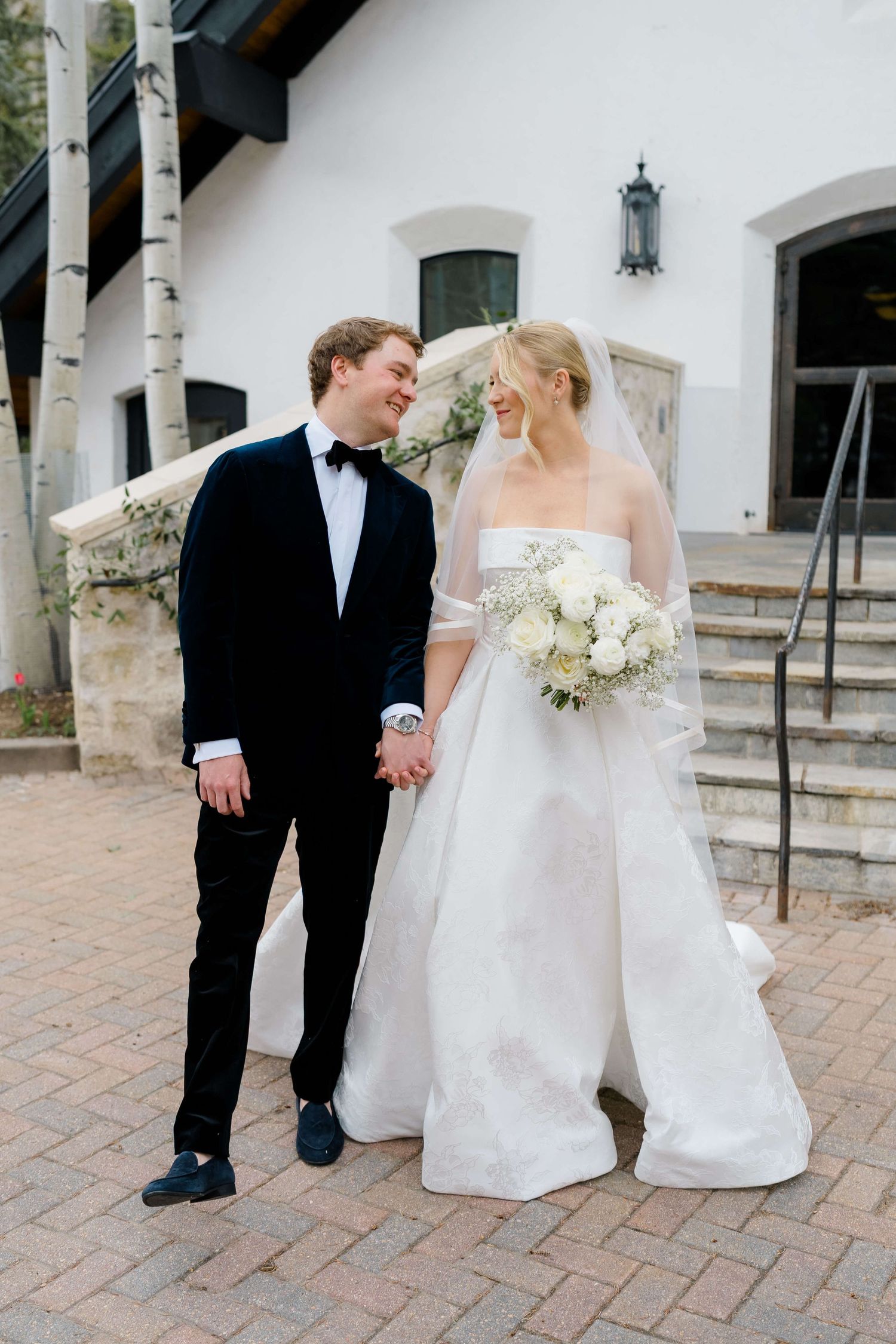 Bride in white strapless gown holding bouquet stands with groom in black tuxedo outside white building.