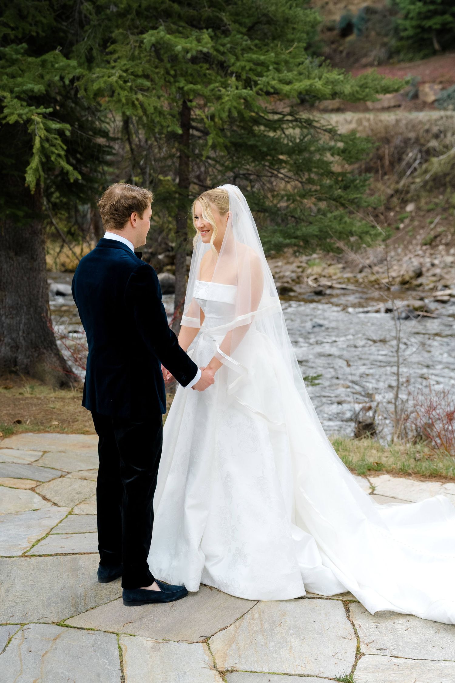 Bride and groom holding hands outdoors near a river, wearing white gown with veil and black tuxedo on stone patio.