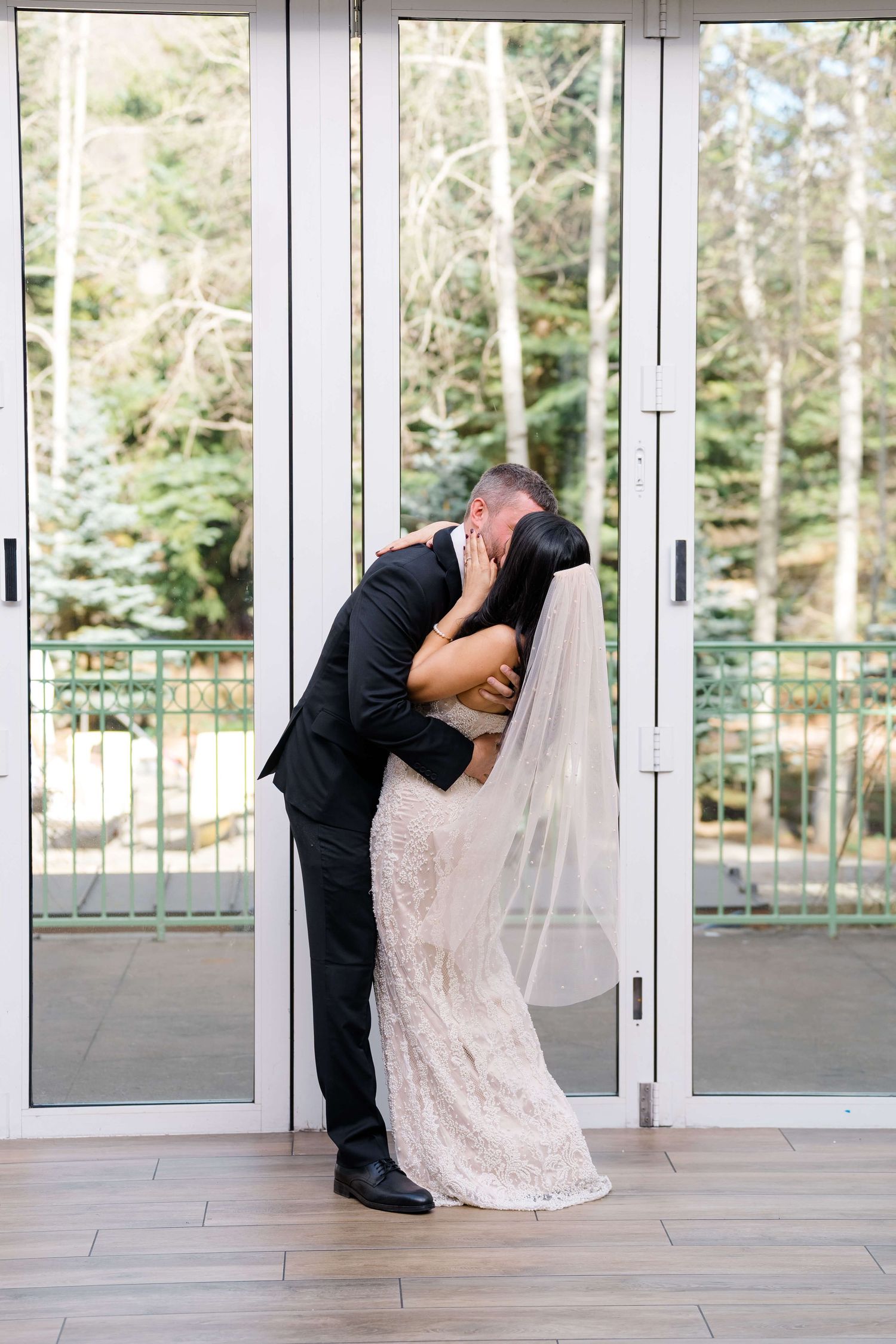 Bride and groom sharing a romantic dip kiss in front of glass doors, bride in lace gown with veil.
