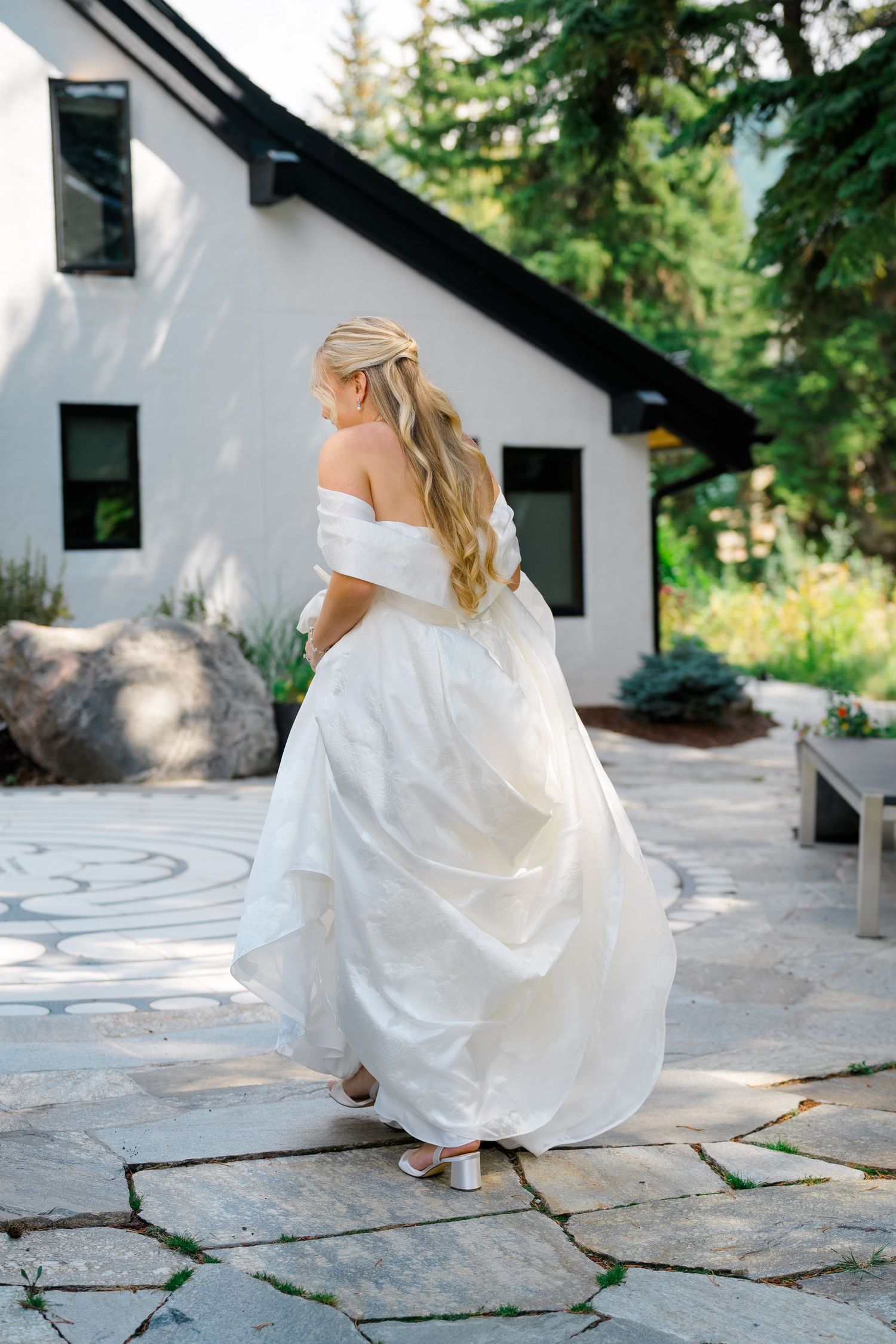 A bride in a flowing white off-shoulder gown twirls on a stone patio outside a modern white house with black trim during vail wedding.
