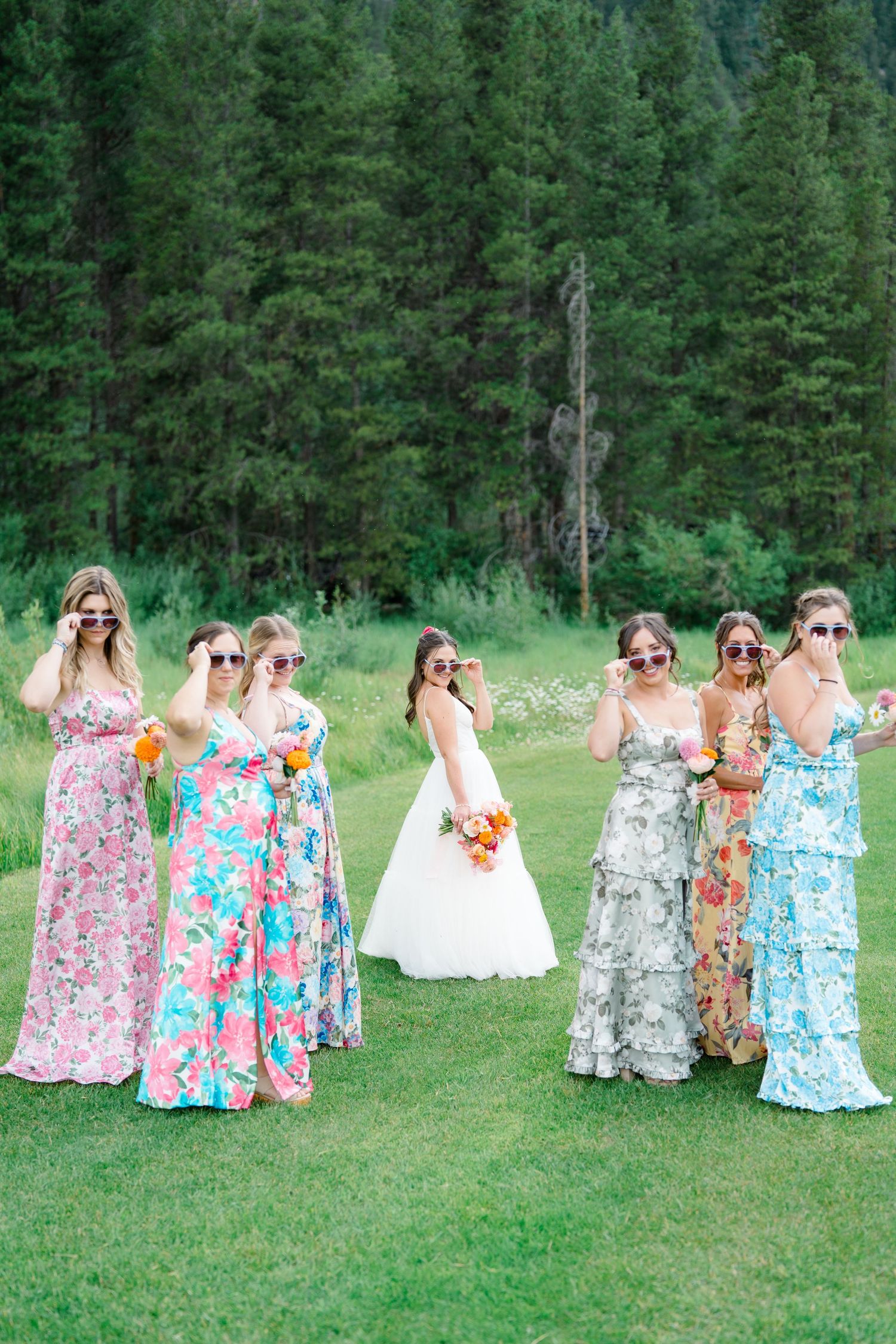 Wedding party in floral dresses laughing together on a grassy field with forest backdrop.