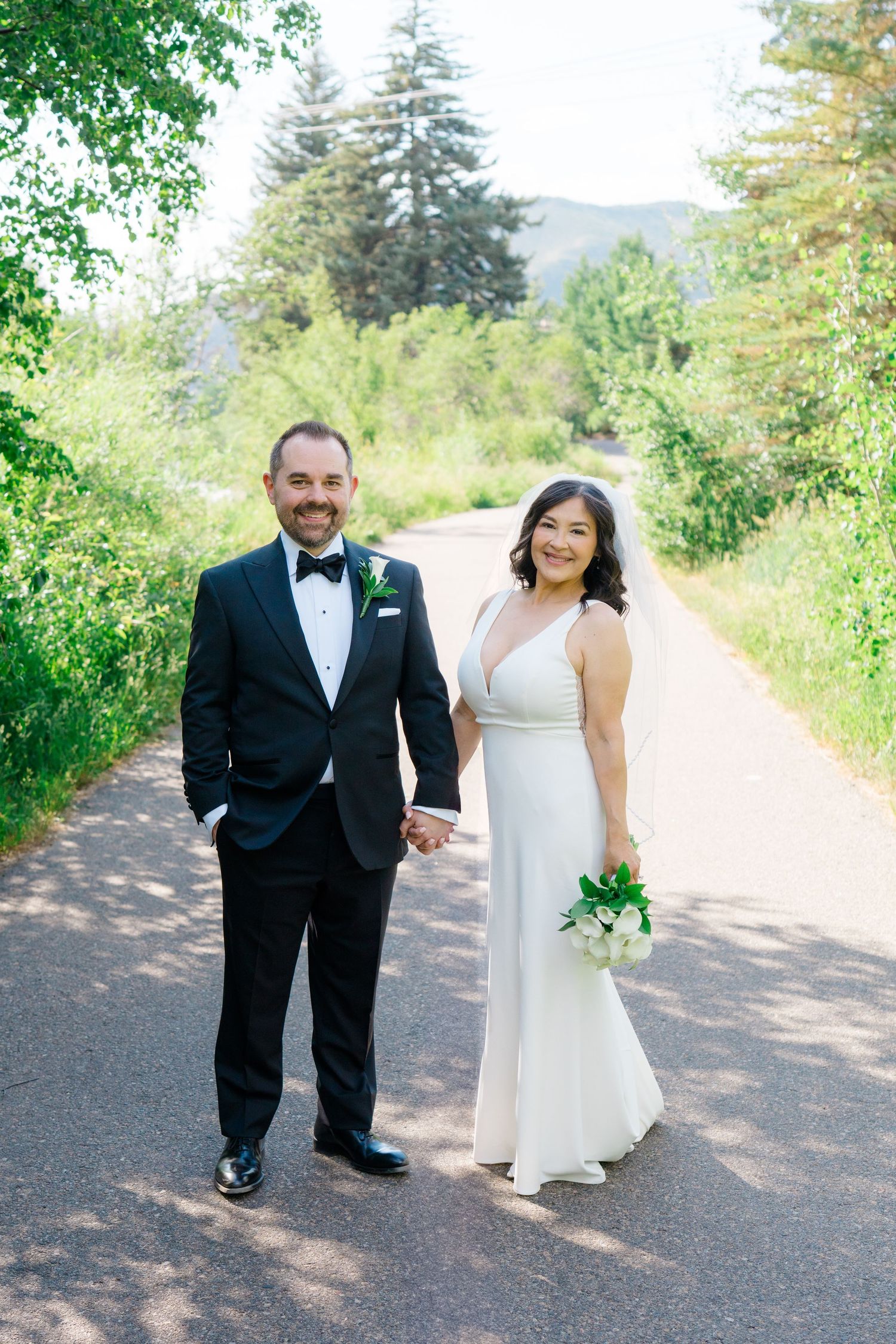 A couple in wedding attire stands on a tree-lined path in a scenic mountain setting.