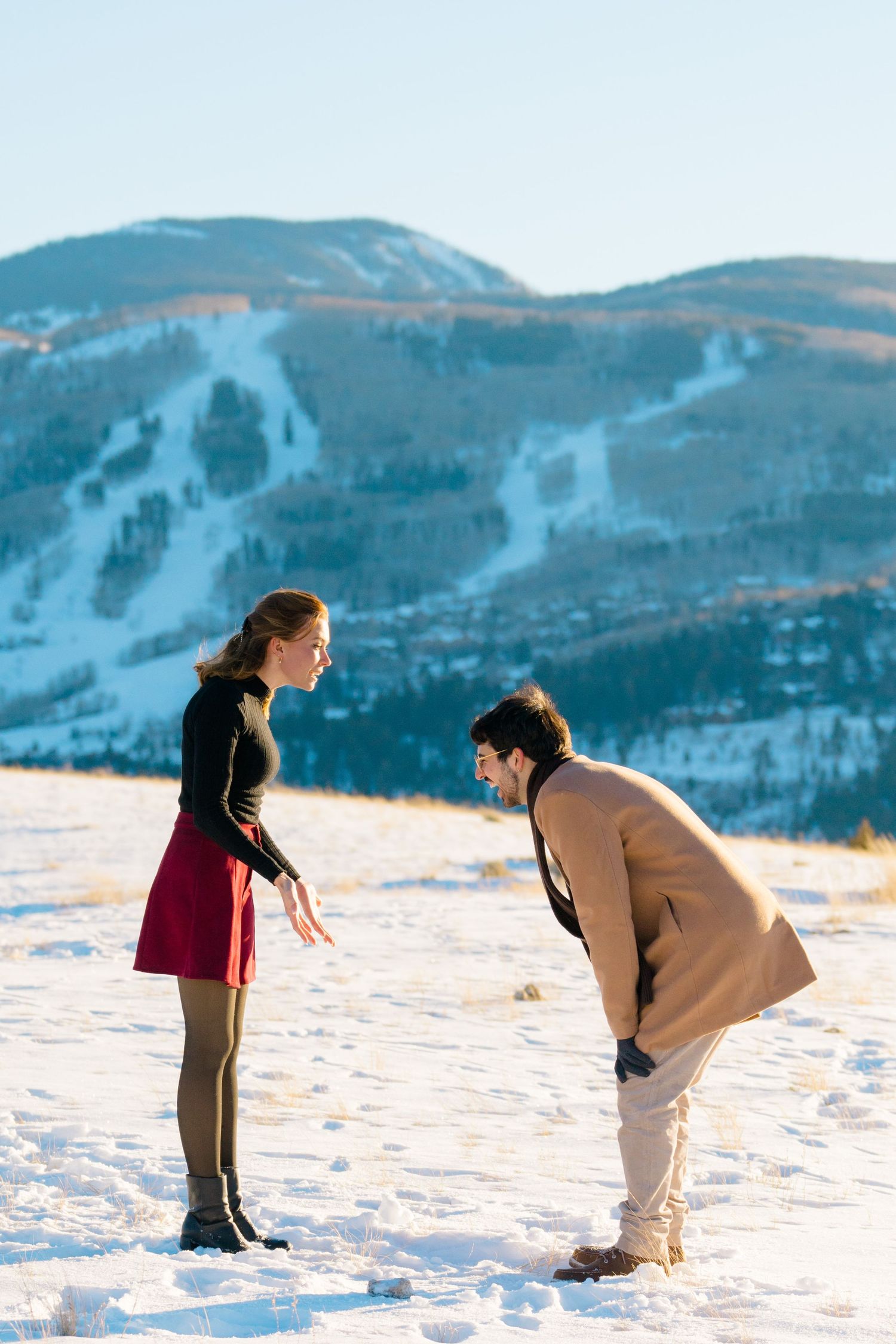 A couple plays in the snow with snowy mountain slopes visible in the background during a winter engagement photoshoot.