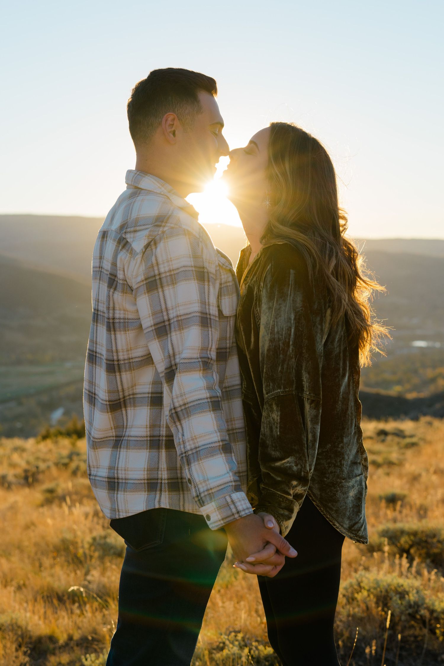 Couple stands in golden sunlight on hillside at sunset wearing plaid shirt and leather jacket.