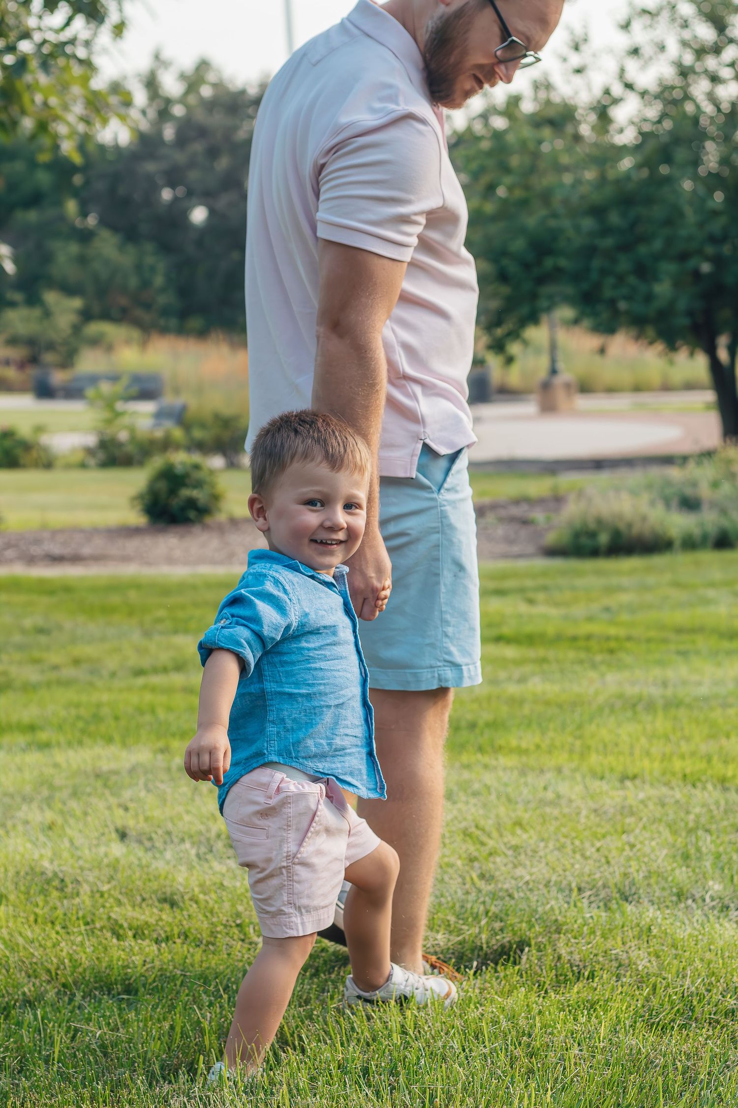 A toddler in a blue shirt and light shorts plays on a grassy lawn during a sunny summer day.