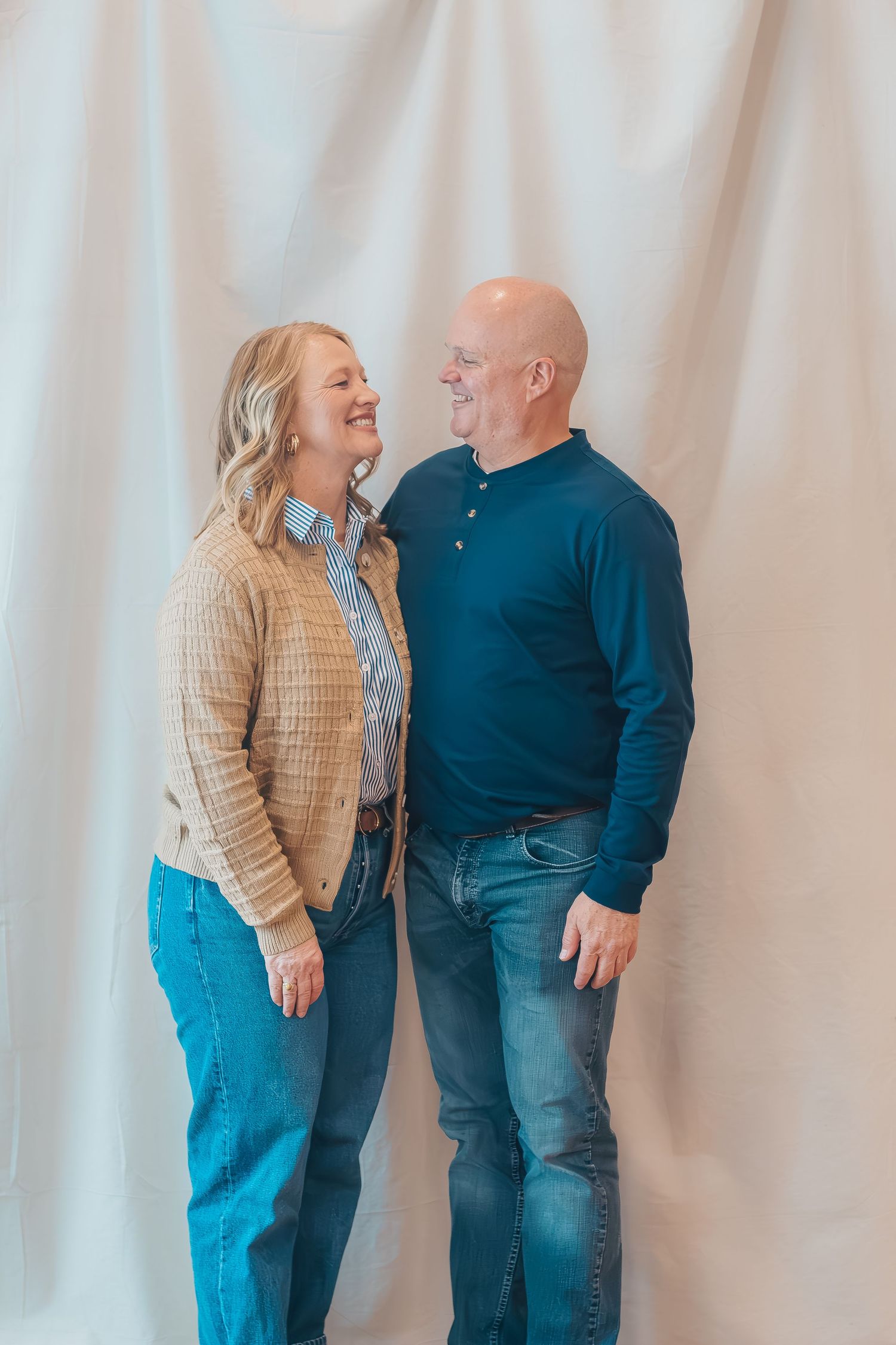 A couple in casual attire laughs together against a white backdrop in a series of candid poses.