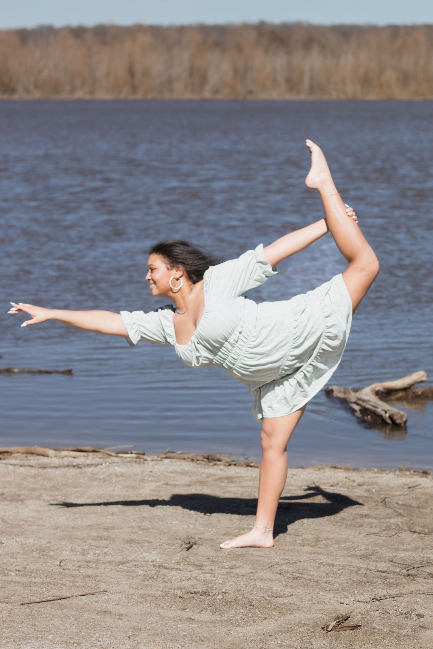 A dancer performs a graceful horizontal pose on a sandy beach next to a lake.