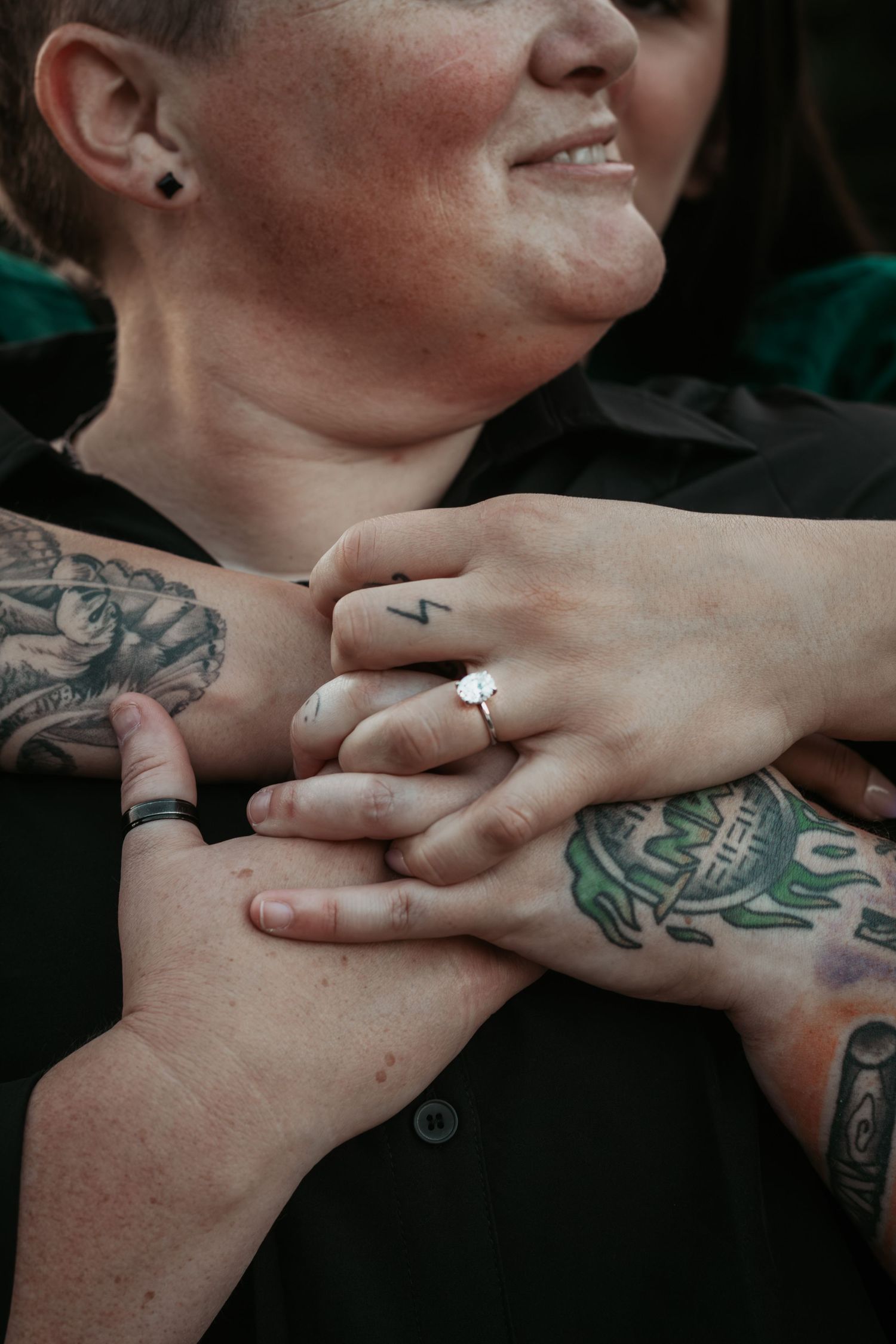 Two tattooed women hold hands, one wearing an engagement ring, smiling warmly in a close romantic moment.