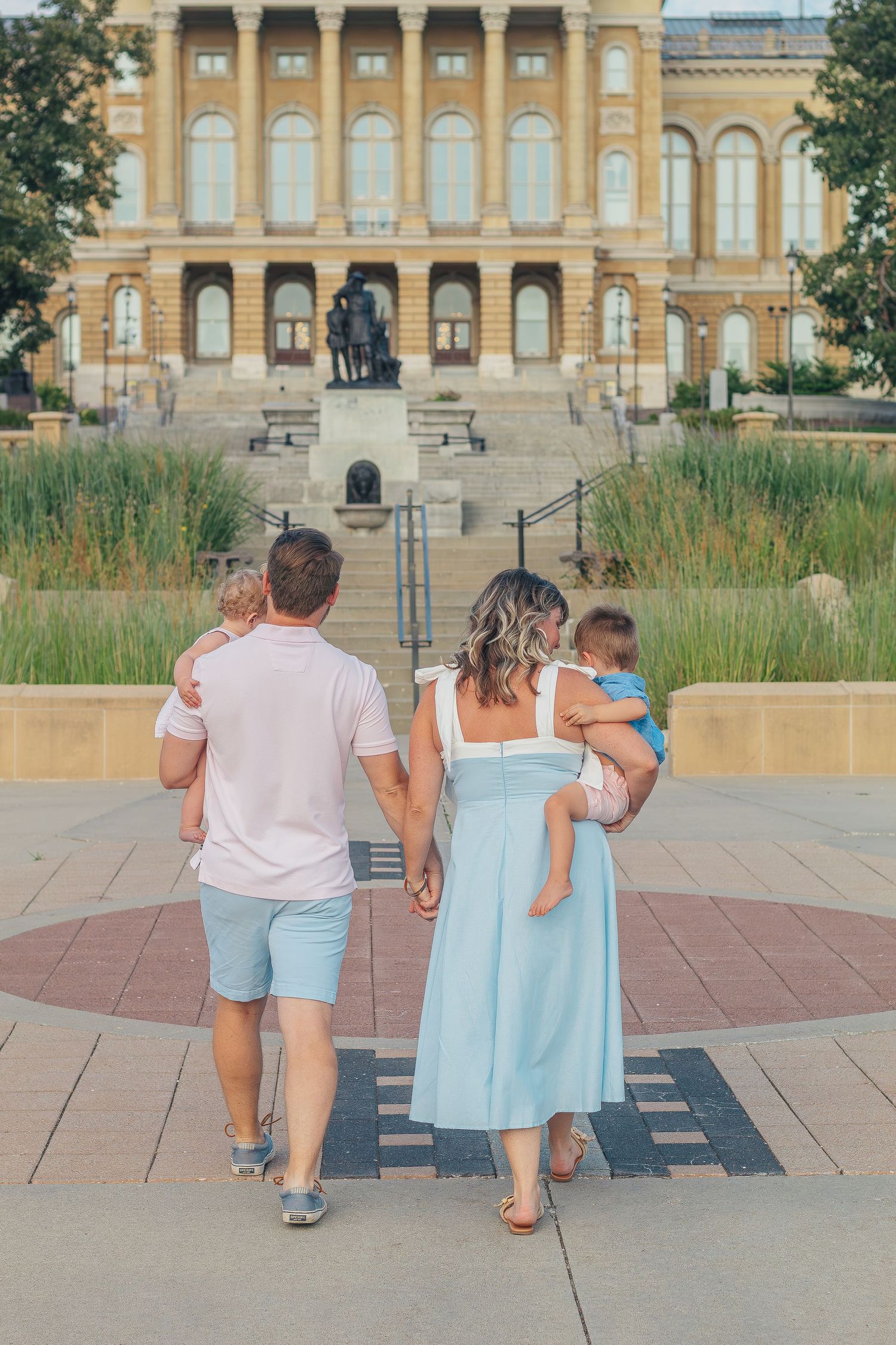 A young family with two toddlers walks toward a grand government building, viewed from behind on a sunny day.