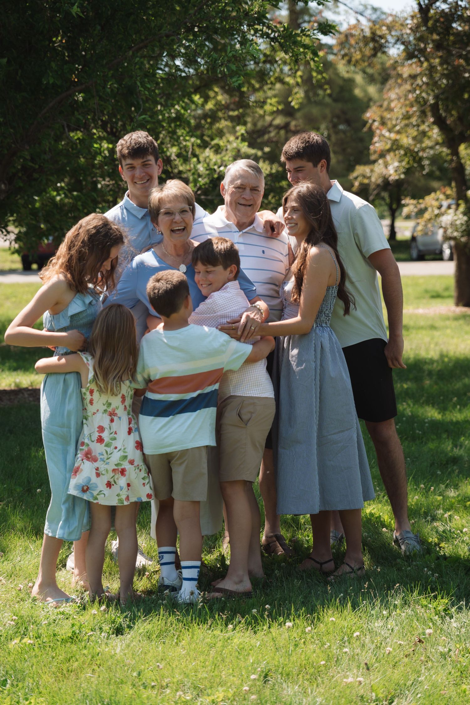 Multi-generational family group hugging outdoors in sunny park, grandparents surrounded by grandchildren smiling together.