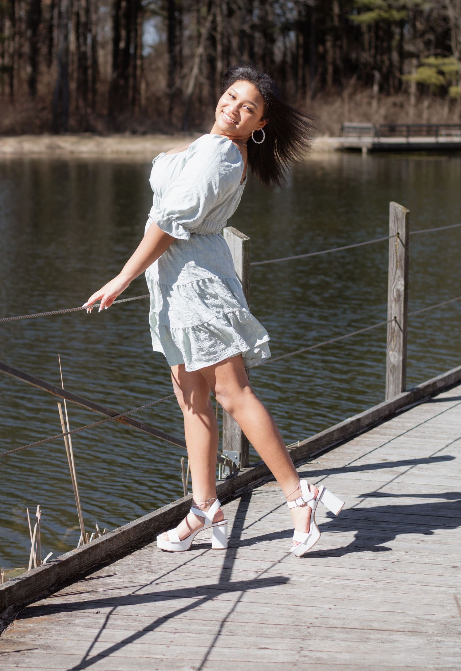 A person in a white dress dances playfully on a wooden dock by a serene lake on a sunny day.