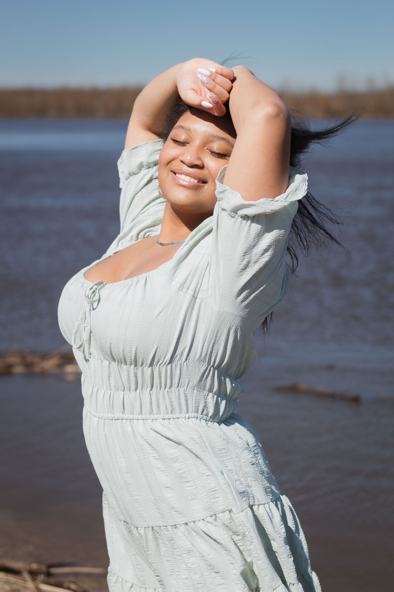 A smiling woman in a light blue dress poses with arms raised near a serene river on a sunny day.