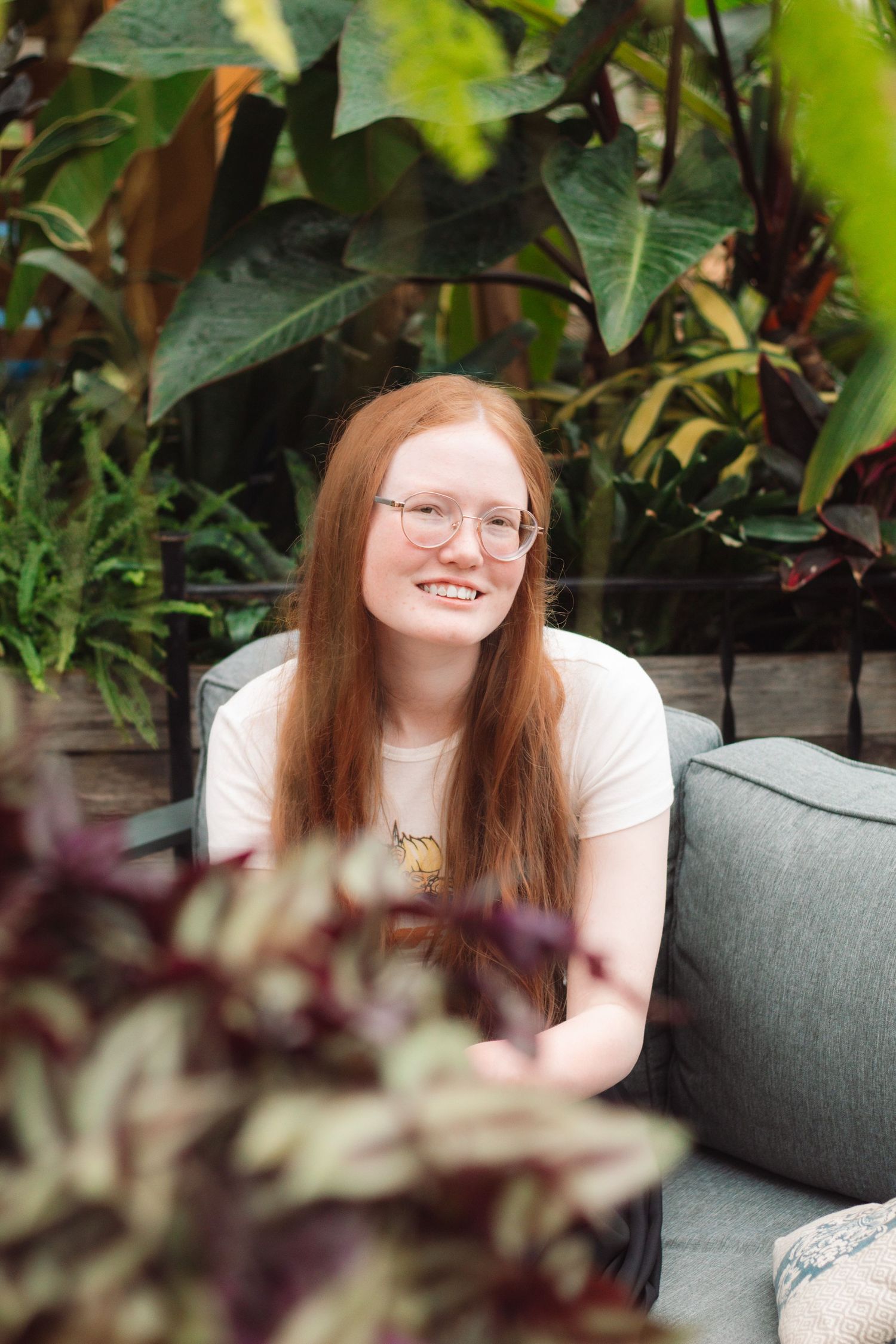 Smiling young woman with long red hair and glasses sitting on a couch surrounded by lush green plants.
