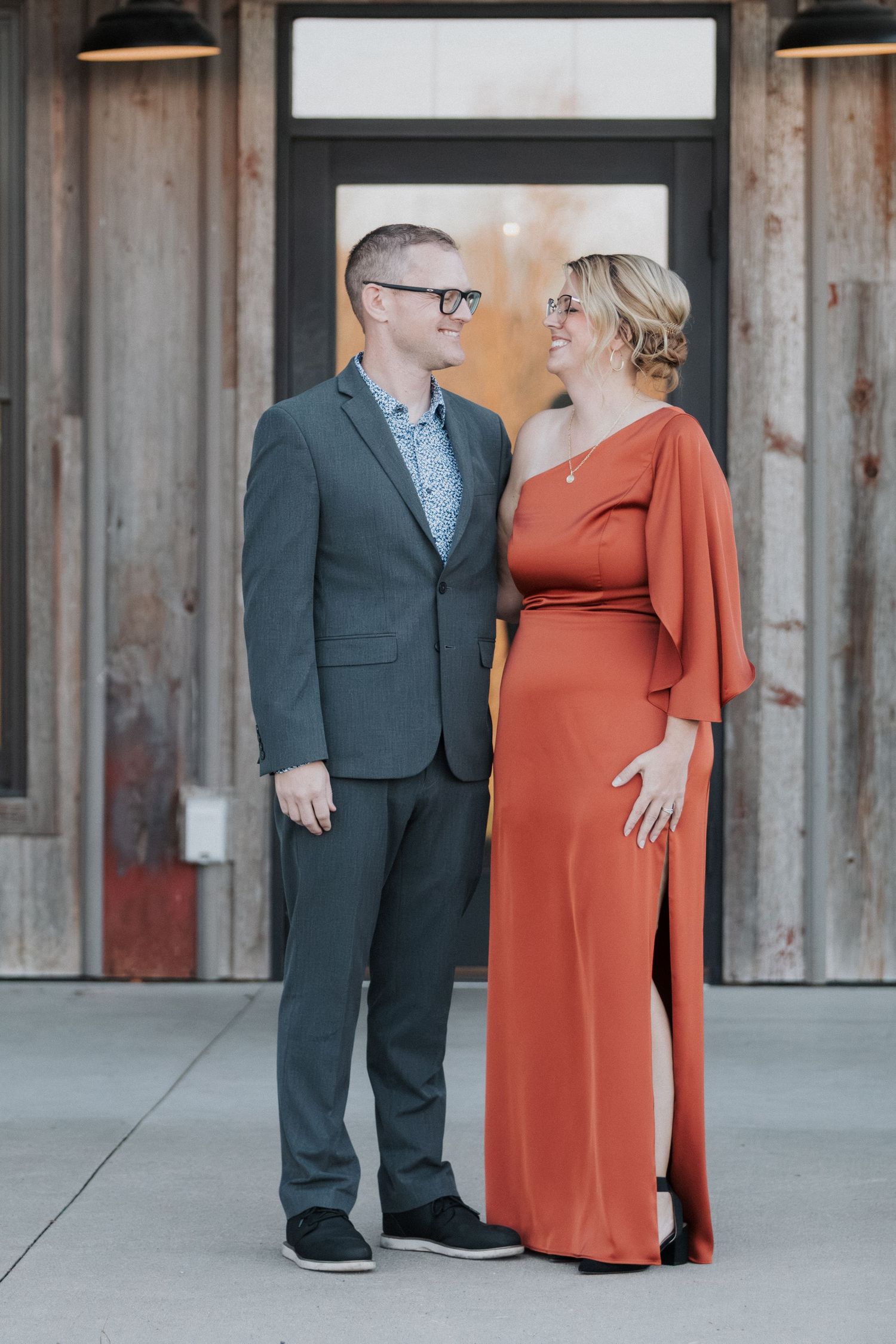 Couple in formal wear, with person wearing gray suit and person in burnt orange dress, posing against rustic wall.