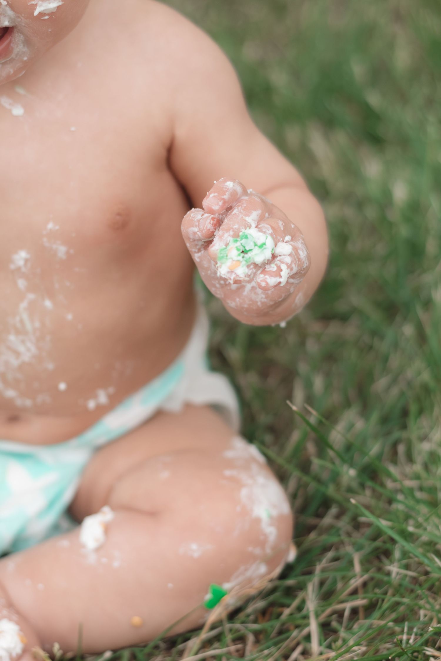 Baby covered in cake frosting sitting on grass during first birthday smash cake celebration outdoors.