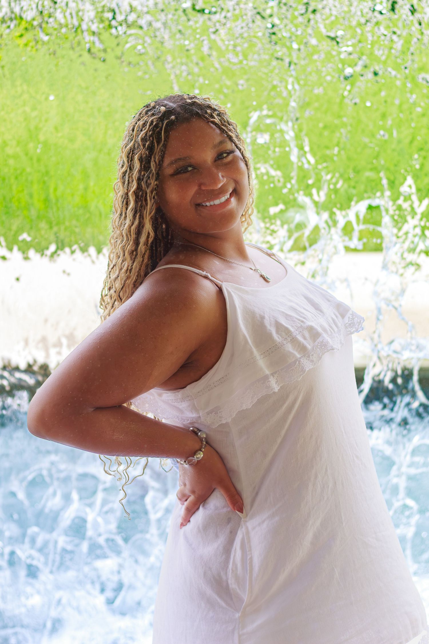 Smiling woman in white dress poses in front of a water fountain with green grass background.
