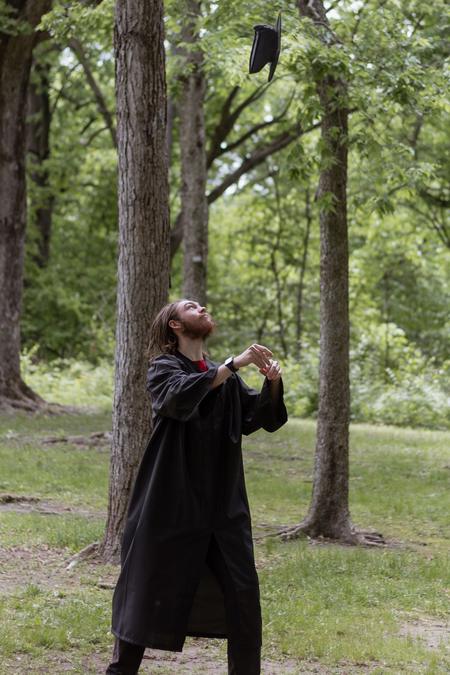 A person wearing a black graduation gown spins joyfully in a wooded outdoor setting during a photoshoot.