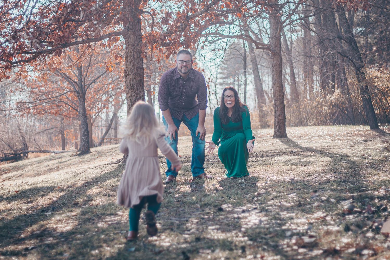 Family photo session in autumn woods with parents and young daughter running toward them on fallen leaf covered ground.