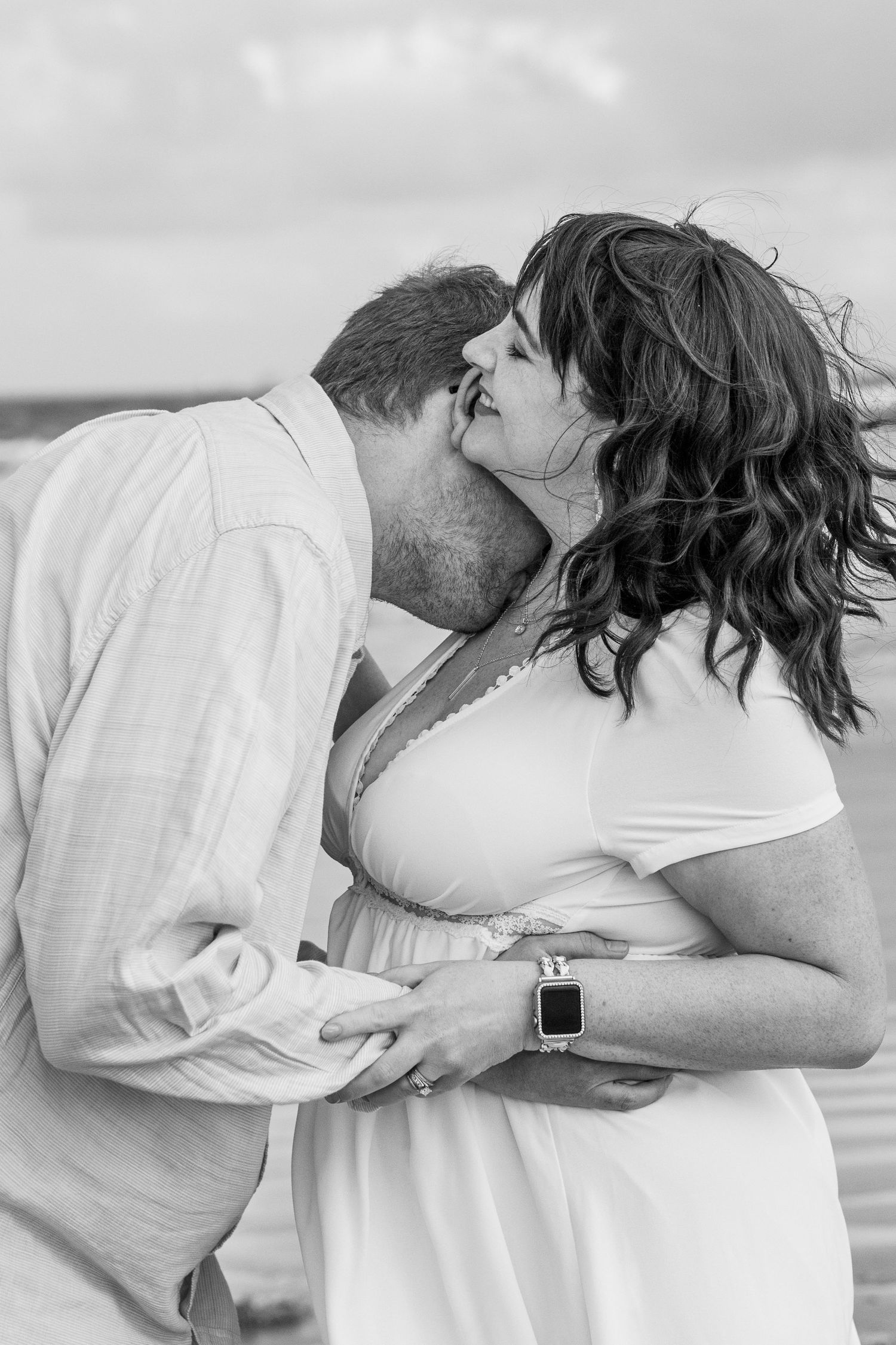 A couple shares an intimate moment in white clothing on a cloudy beach.