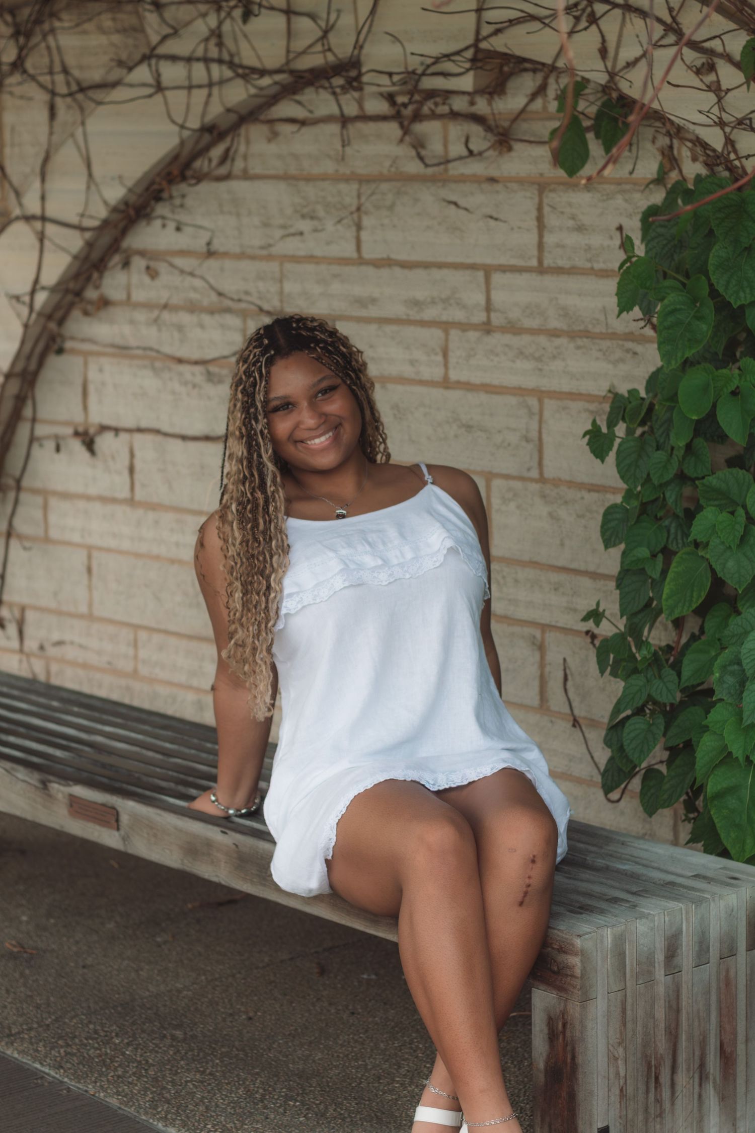 Young woman with braided hair smiling in white sundress, seated on bench with stone wall and green vines backdrop.