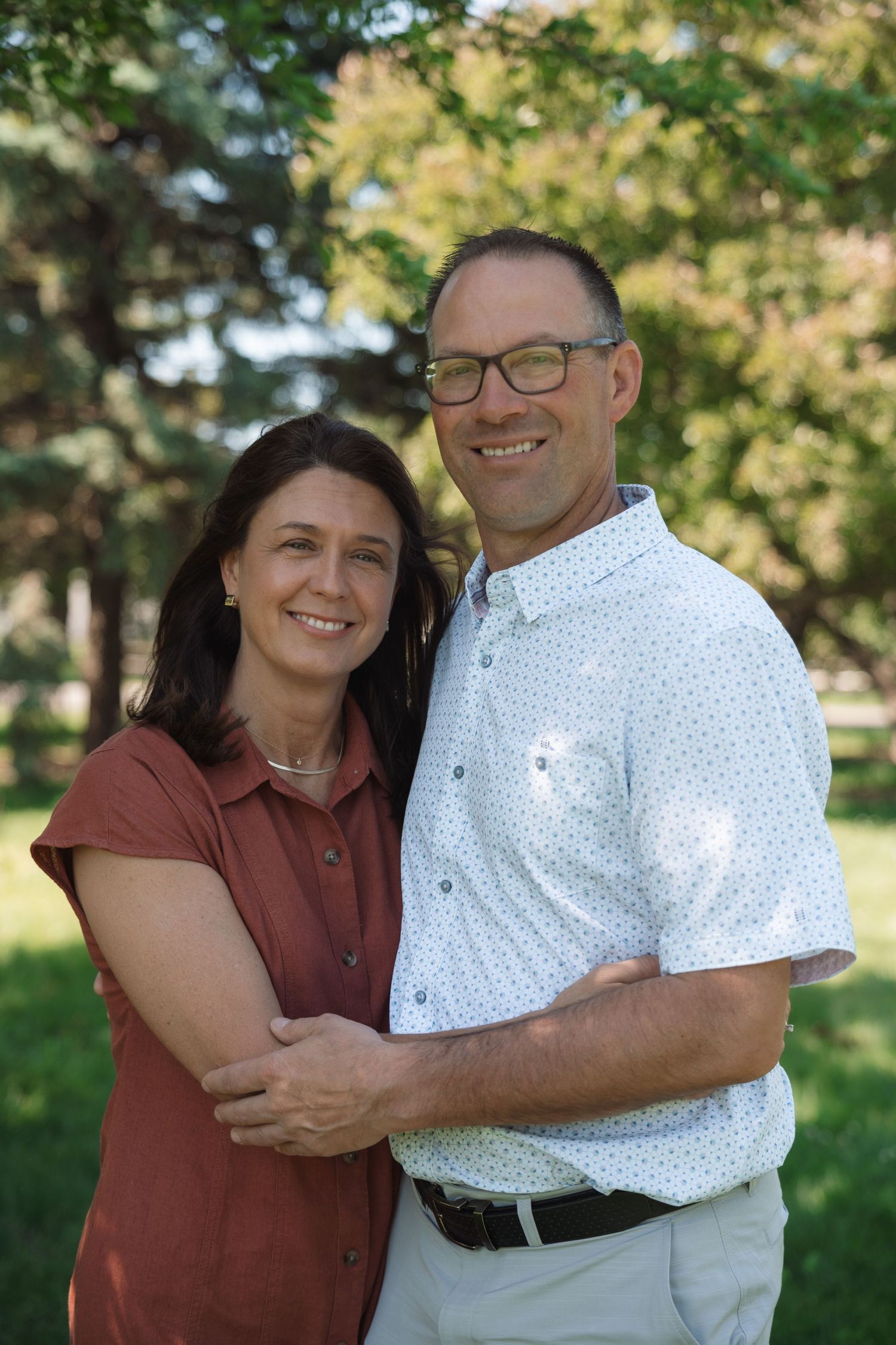 A smiling couple embraces outdoors in a lush green park, the woman in red and man in a white patterned shirt wearing glasses.