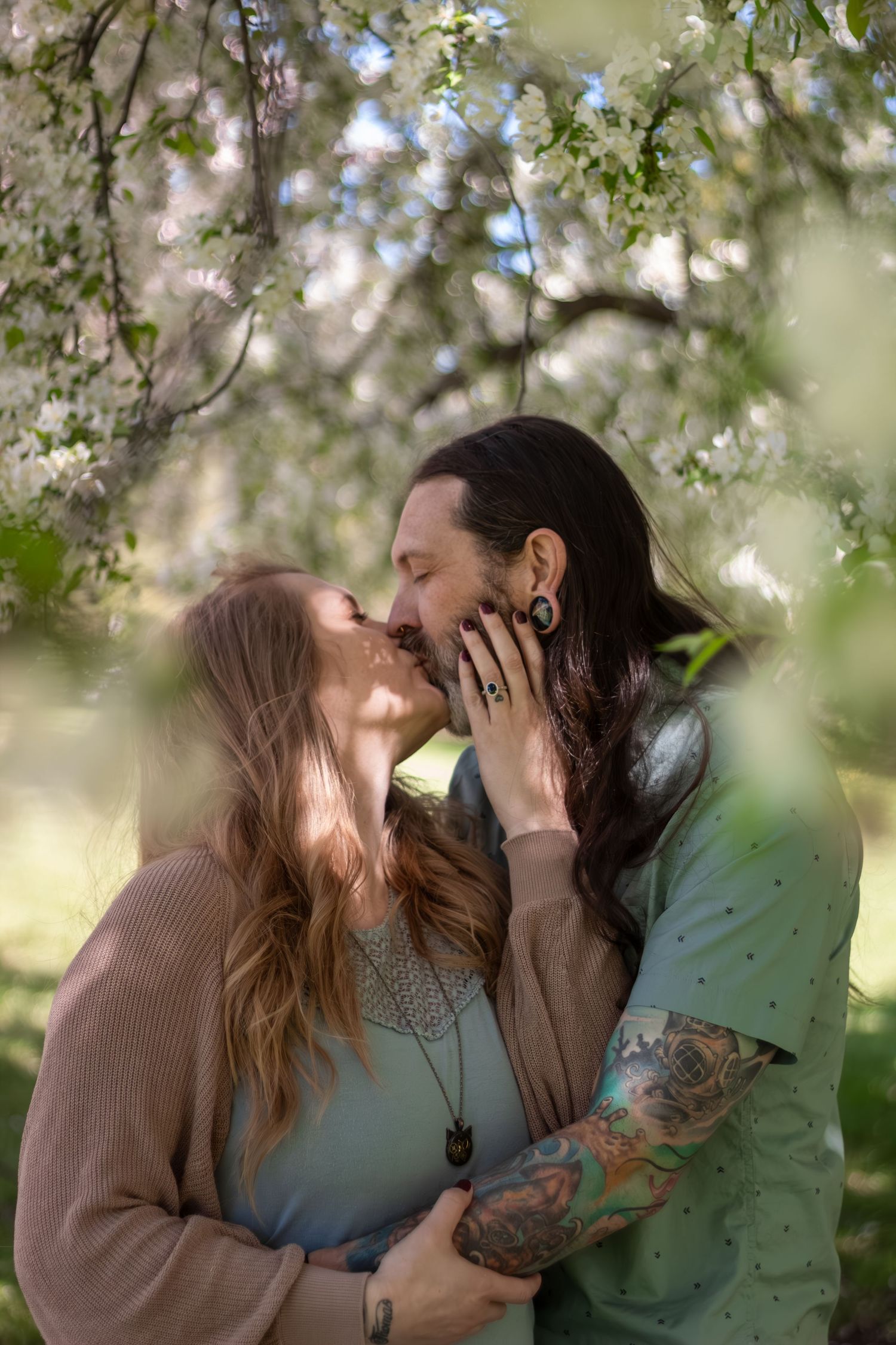 A romantic couple shares tender kisses under blooming spring tree branches in a dreamy outdoor setting.