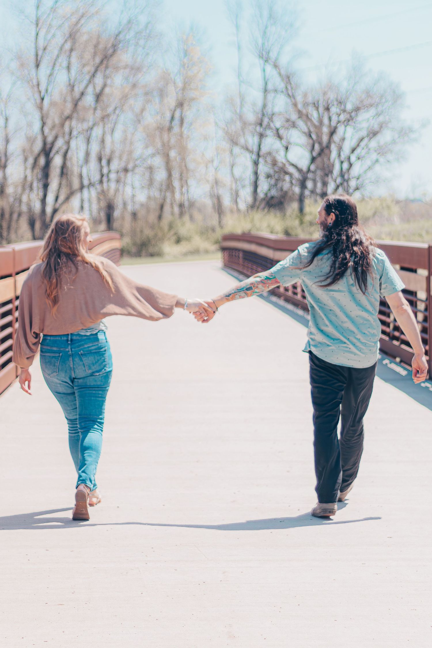 Two people holding hands while walking across a wooden bridge in casual attire on a sunny day.