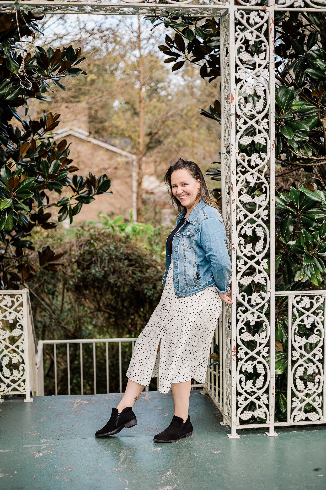 Person in denim jacket and polka dot dress sits on ornate white metal garden bench surrounded by greenery.