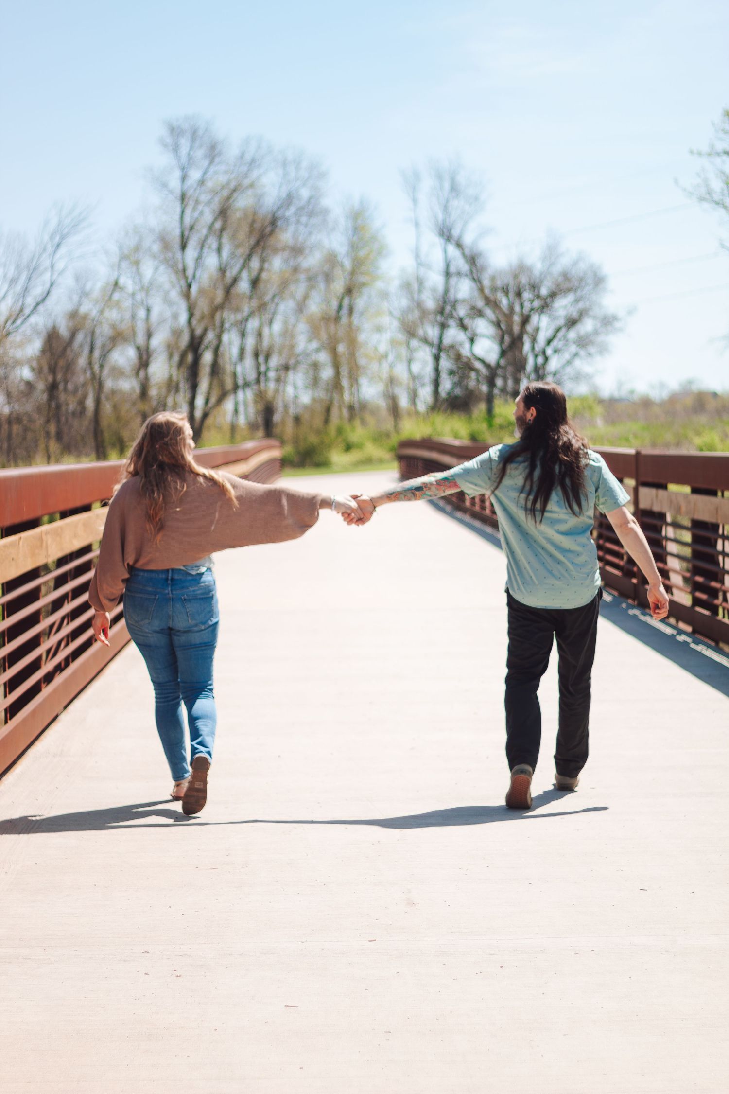 Two people holding hands walk together down a wooden bridge pathway on a sunny spring day.