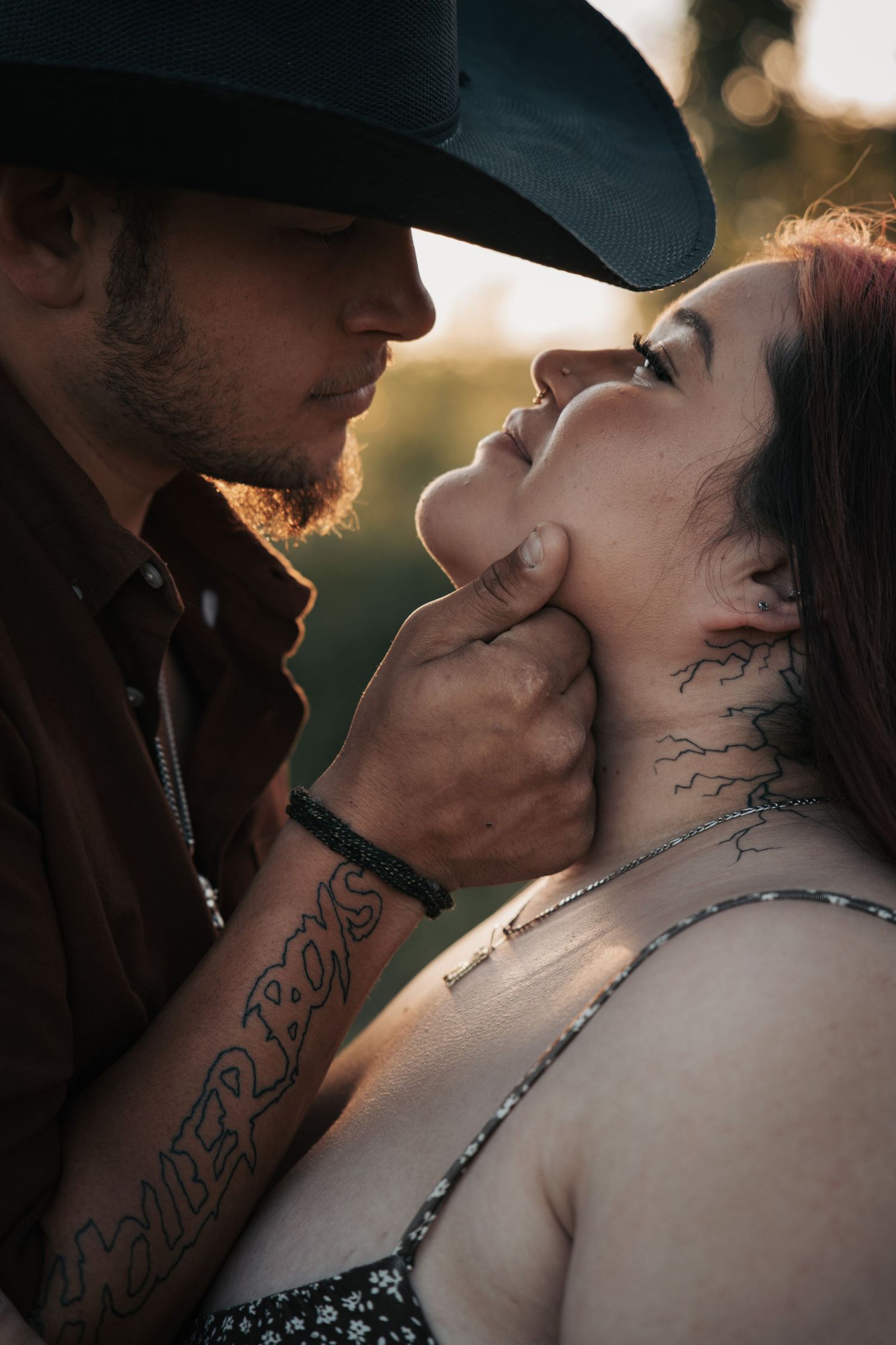 A tattooed couple shares an intimate moment, man in cowboy hat gently holding womans chin outdoors at golden hour.