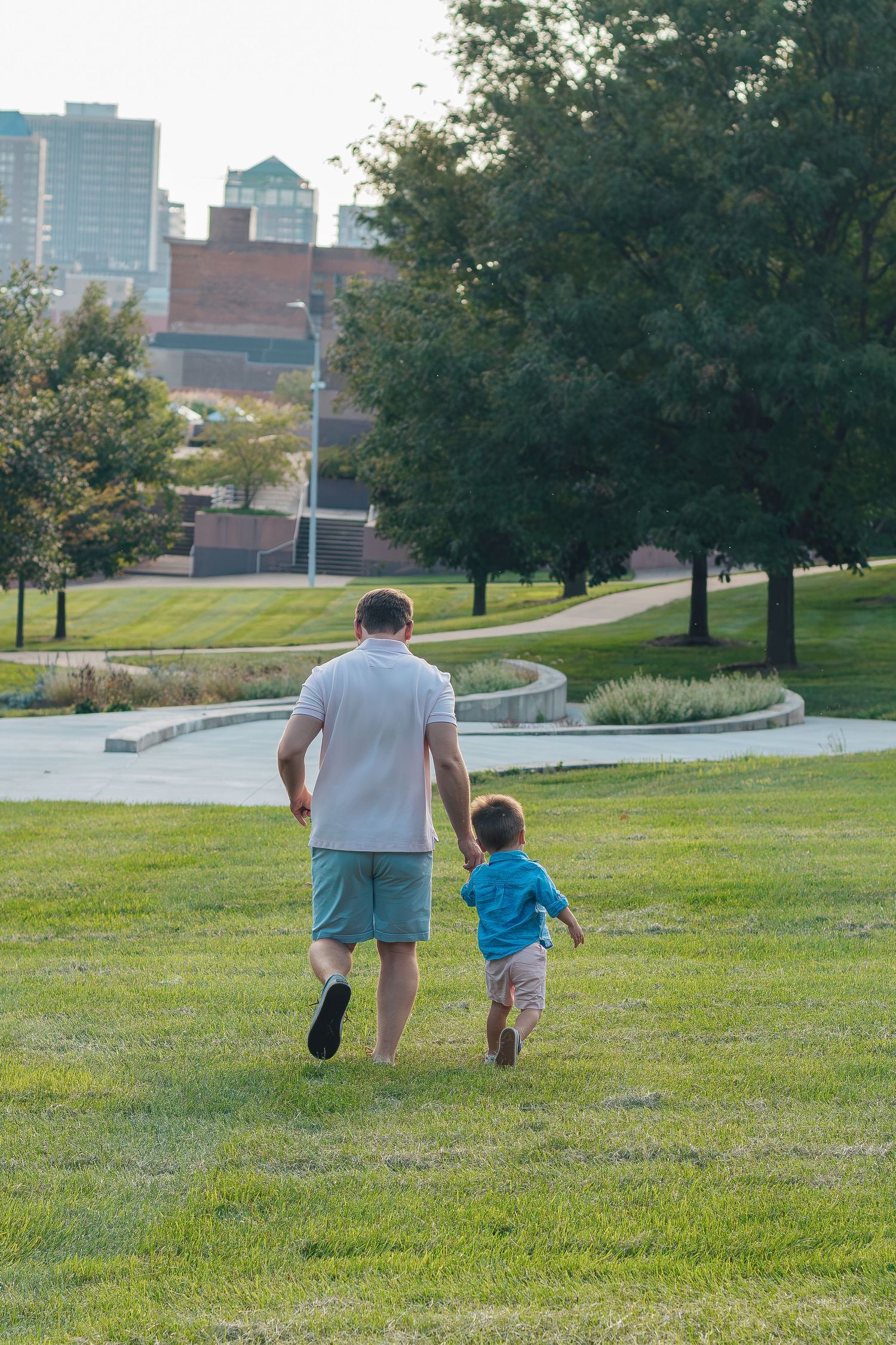 Father and young child walking hand-in-hand across lush green park lawn with city skyline and trees in background.