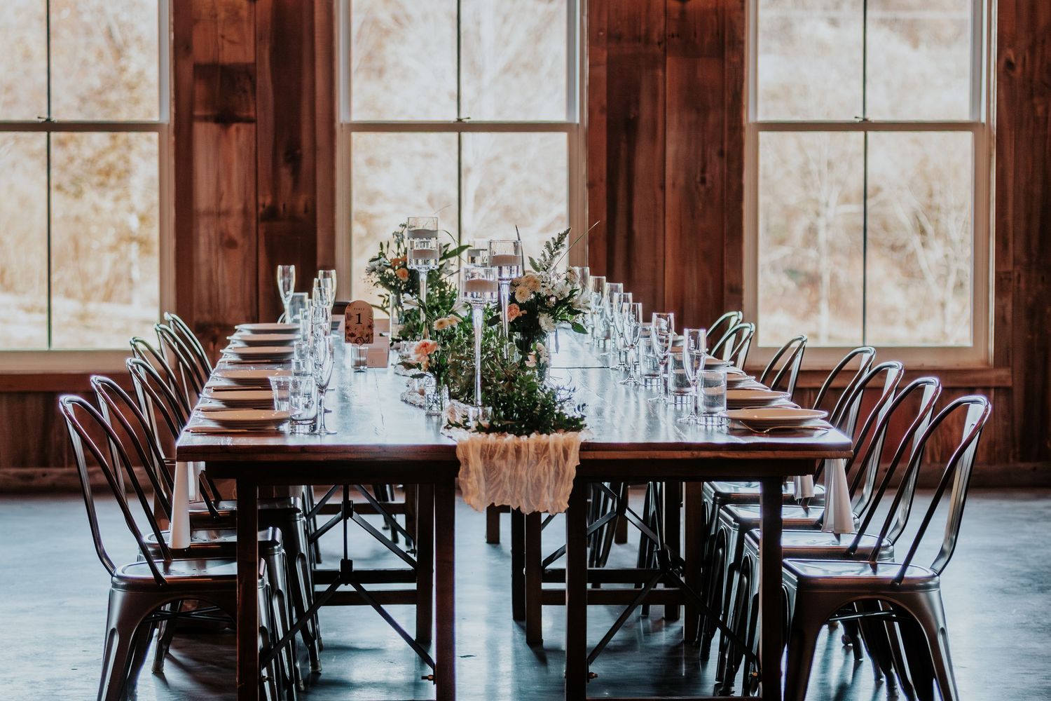 Rustic restaurant interior with wooden tables and chairs arranged in rows by large windows with natural light.