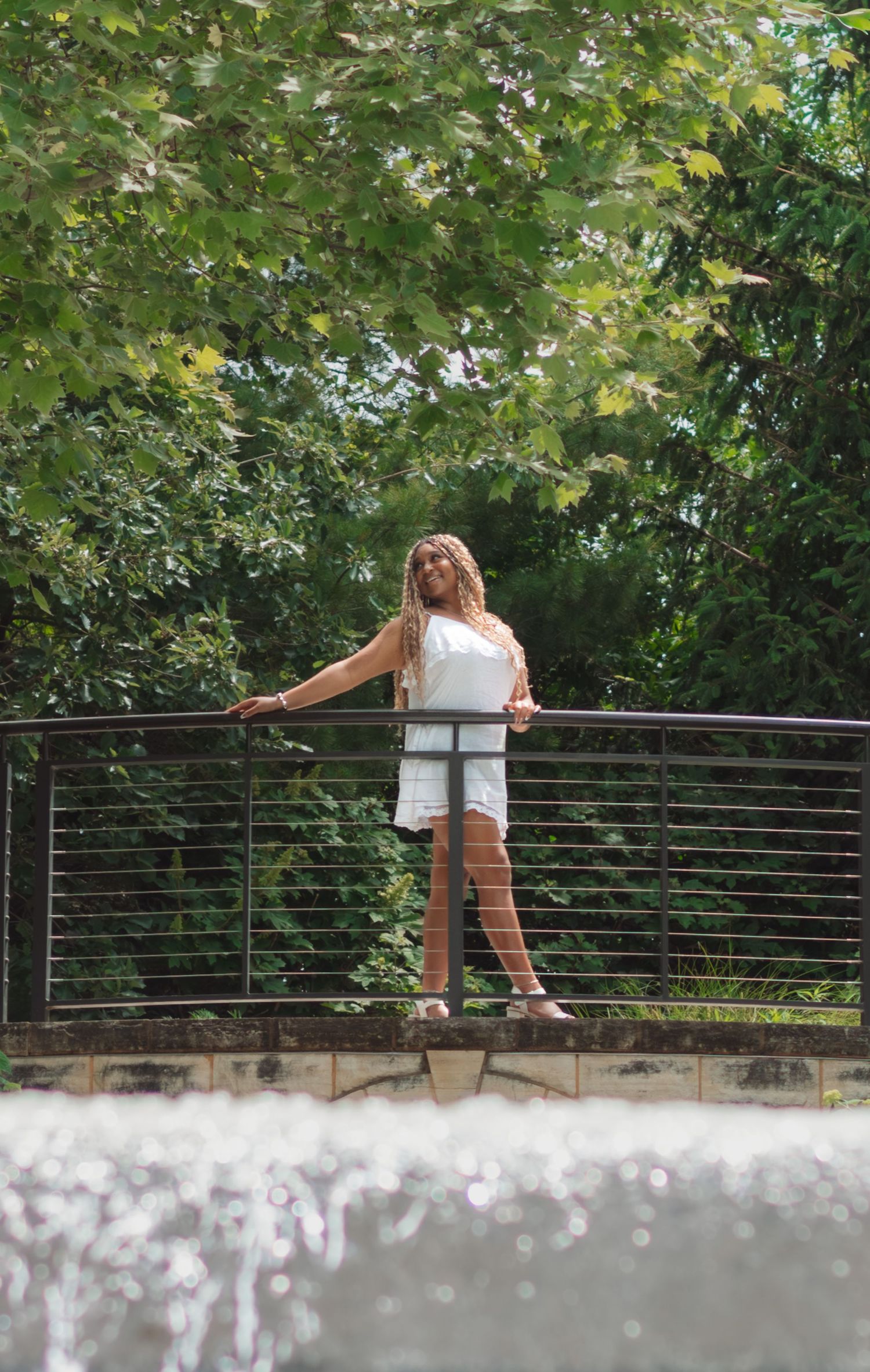 Woman in white outfit standing on a bridge railing surrounded by lush green trees in a park setting.