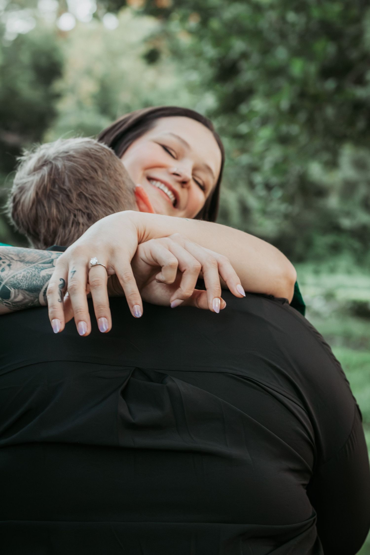 Smiling woman with ring hugs partner outdoors, showing engagement ring on hand with green trees in background.