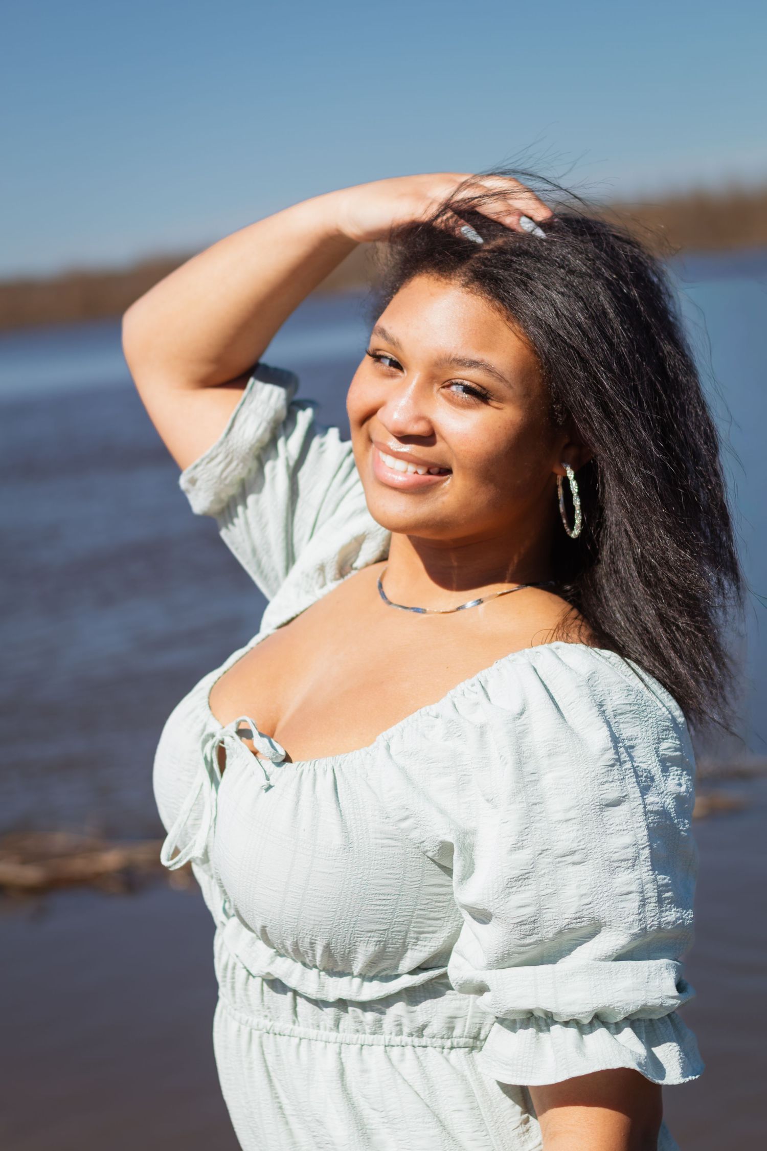 A person in a white ruffled dress poses on a beach with sunlight creating a warm glow.
