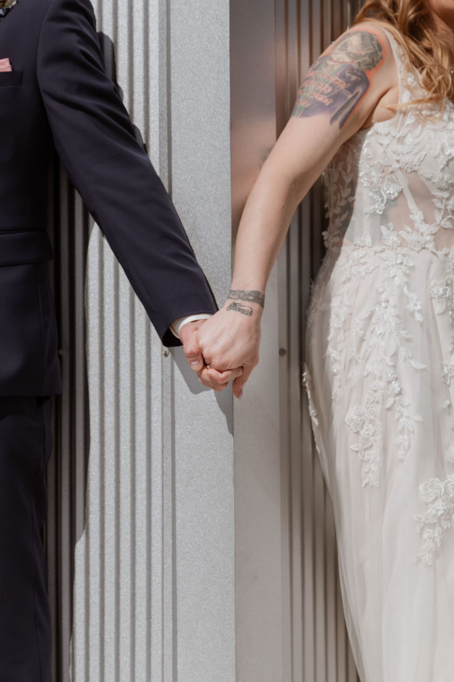 A bride in a lace gown and tattooed groom in navy suit hold hands beside elegant stone columns.
