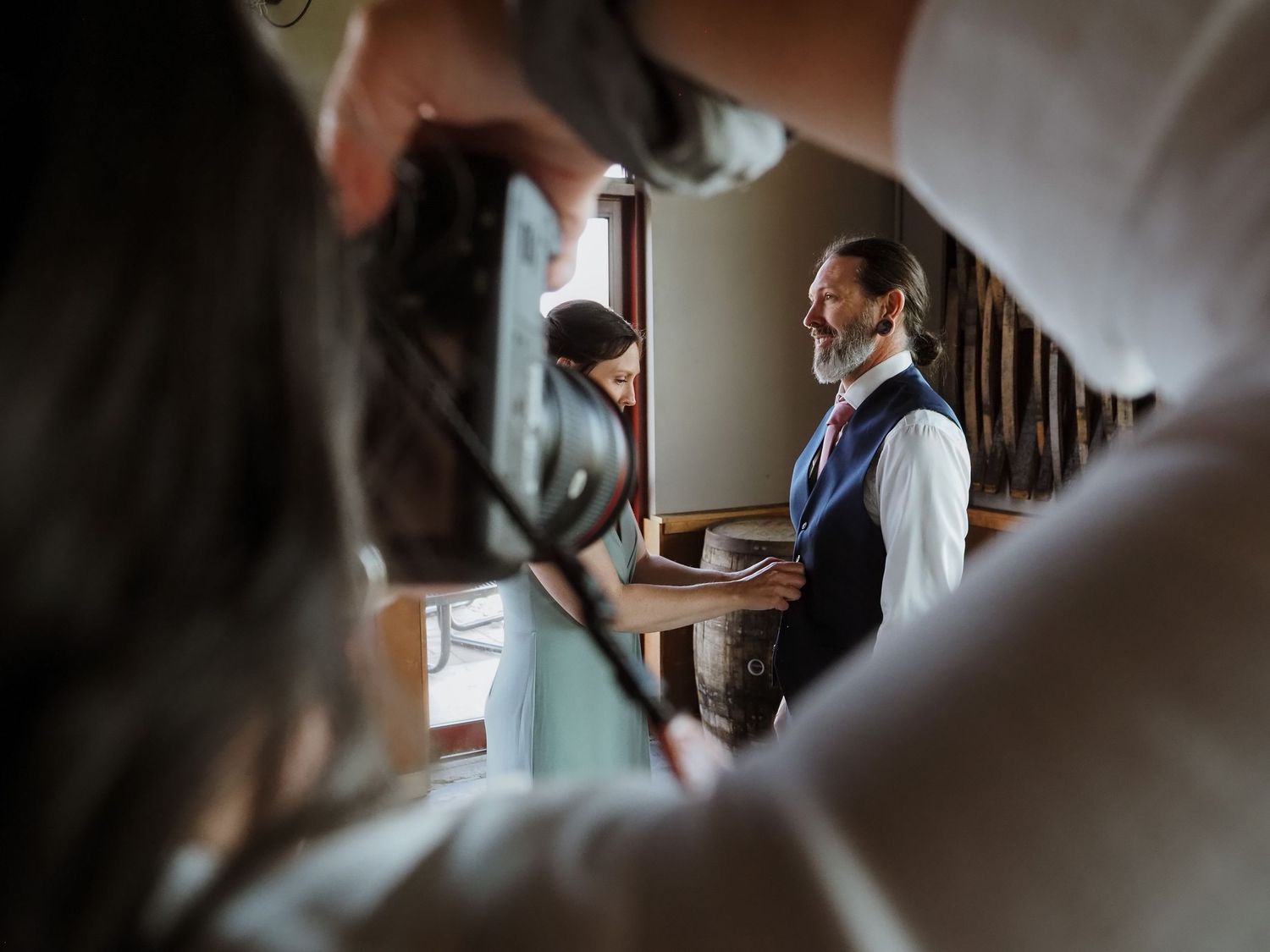 Groom in vest and tie smiles during wedding preparations while surrounded by attendants in a warmly lit room.