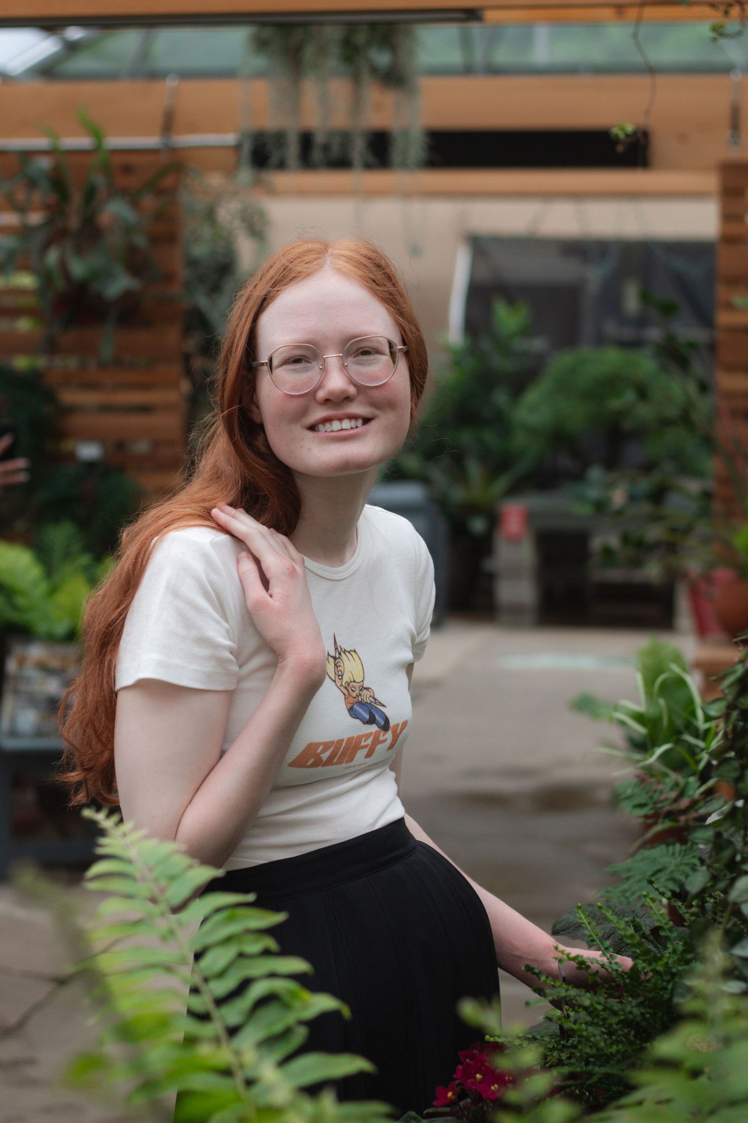 Smiling young woman with long red hair and glasses sits among lush green plants in a greenhouse, wearing a white graphic tee.