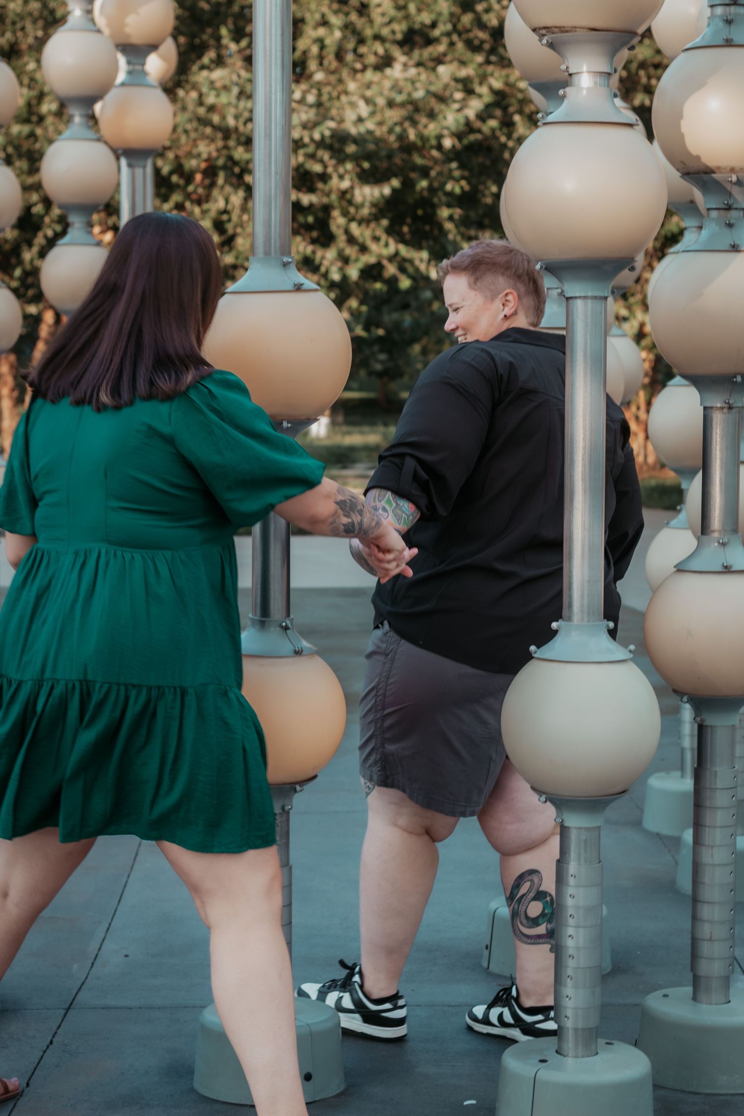 Two people holding hands among decorative globe light posts, one in a green dress and one in a black hoodie with tattoos.