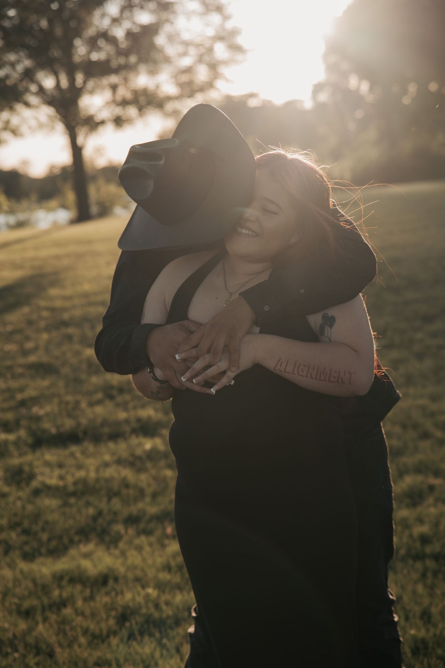 Couple embracing at golden hour in a park, woman laughing joyfully as man in black hat hugs her from behind.