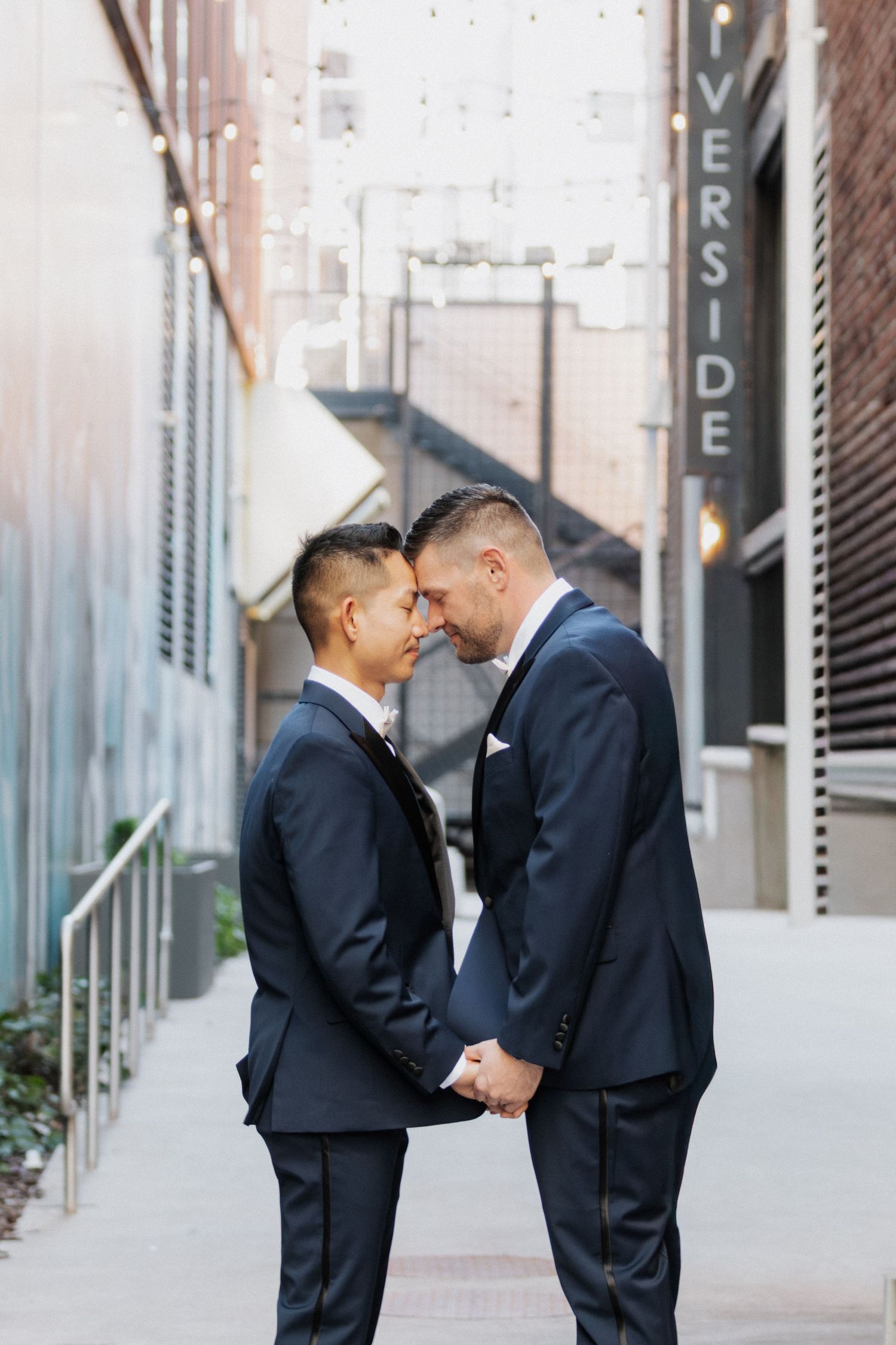A romantic moment between two people in matching navy suits standing close together in an urban alleyway.