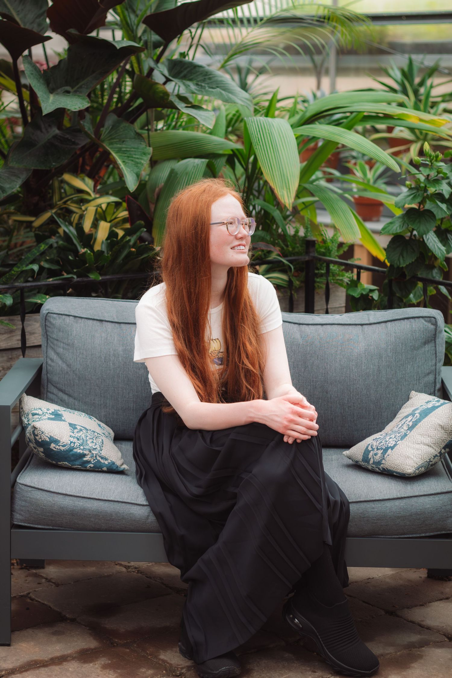 Young redhead woman with glasses sits smiling on gray outdoor sofa surrounded by lush tropical green plants.
