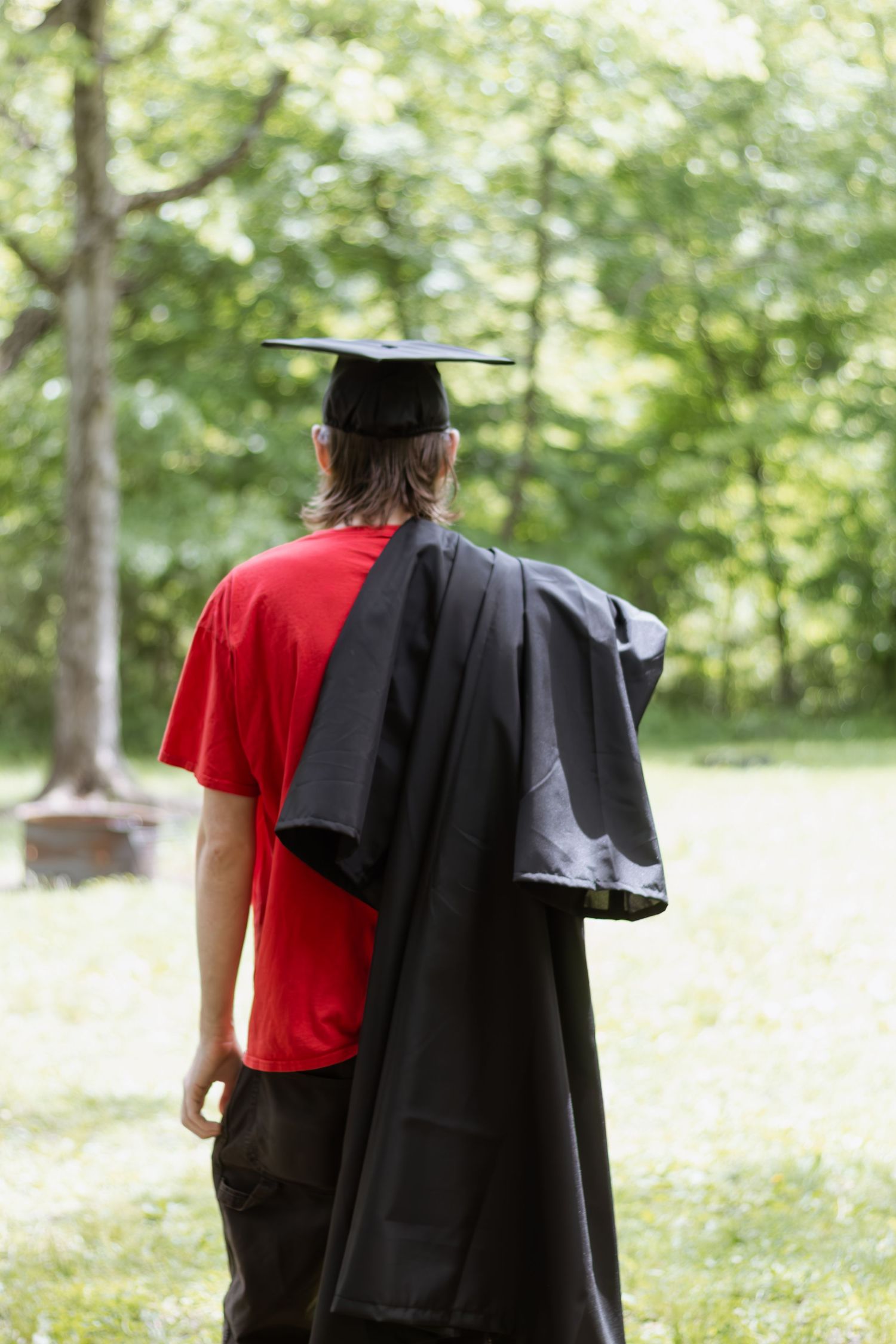 Graduate in black cap and gown with red shirt walks through a sunlit wooded area during commencement ceremony.
