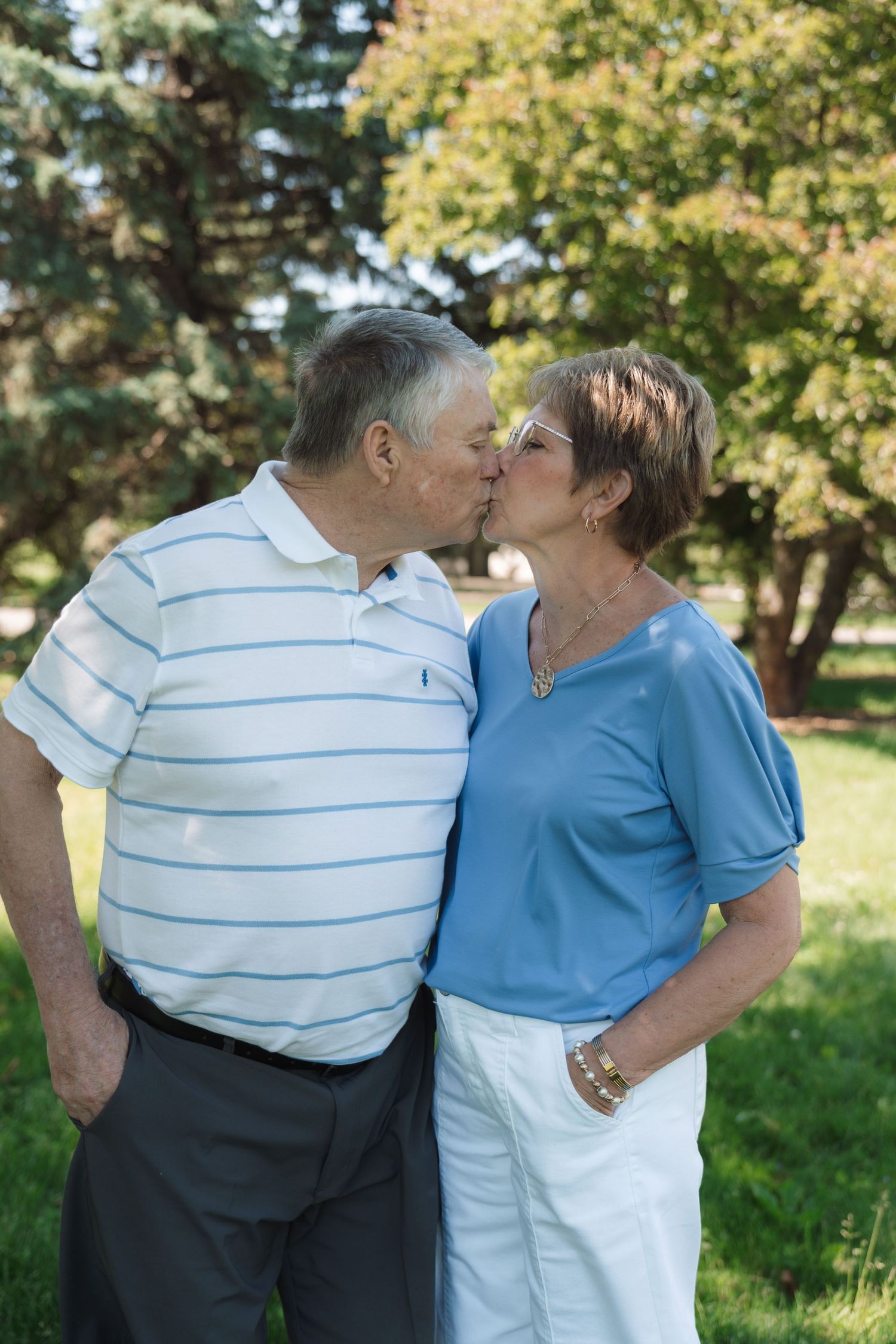 An elderly couple shares a romantic kiss outdoors in a lush green park, dressed in matching blue tops and white bottoms.