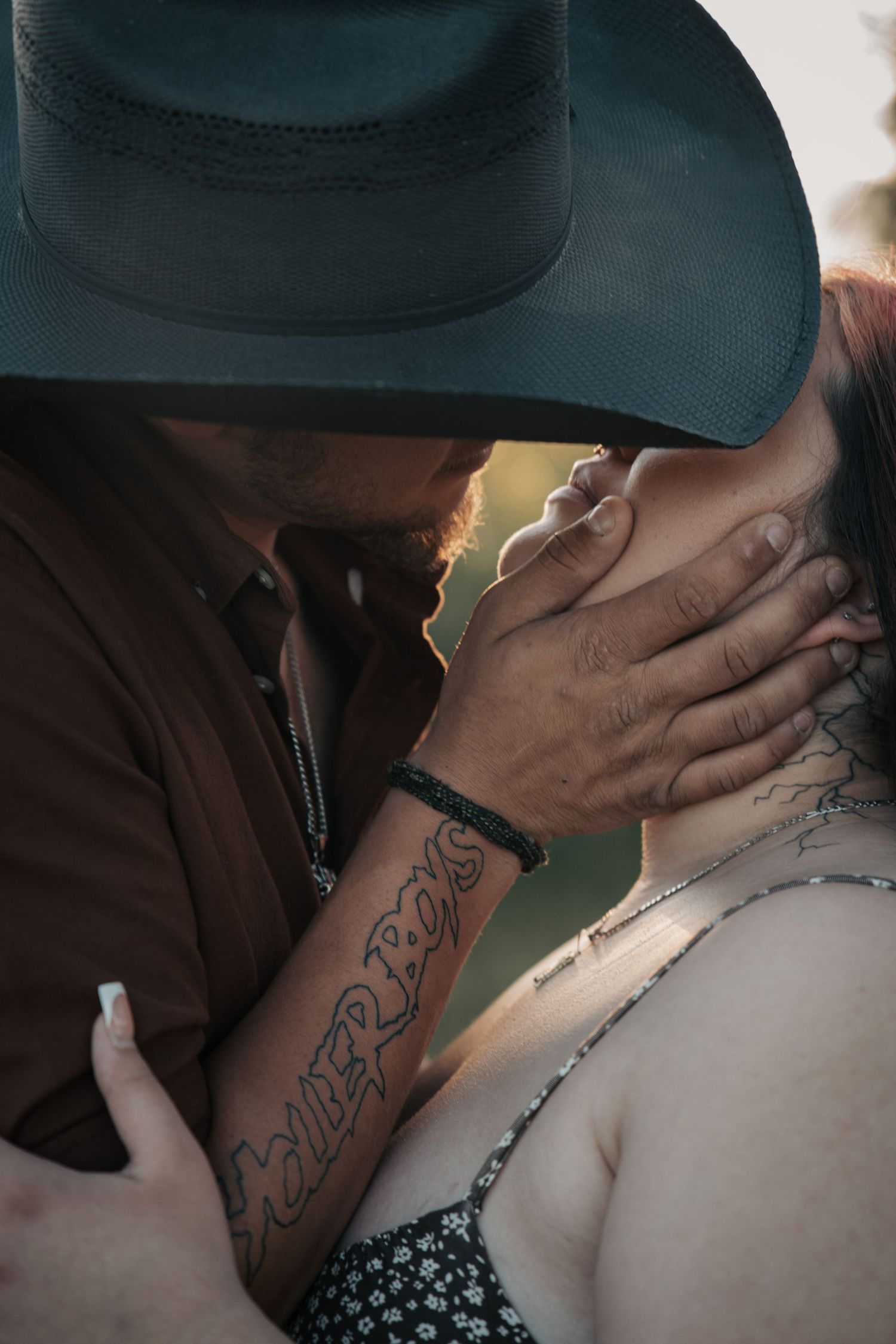 A tattooed man in a black cowboy hat tenderly kisses a woman while cradling her face with his hand.
