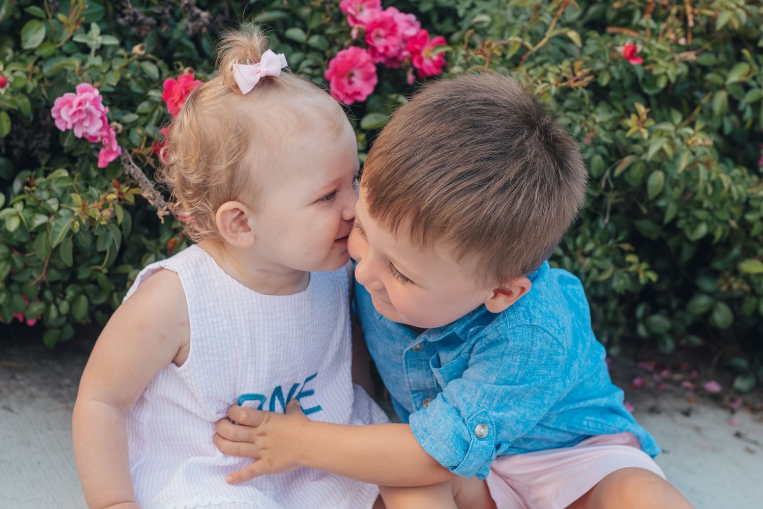 Two young children share a sweet moment together in a garden with pink flowers blooming in the background.
