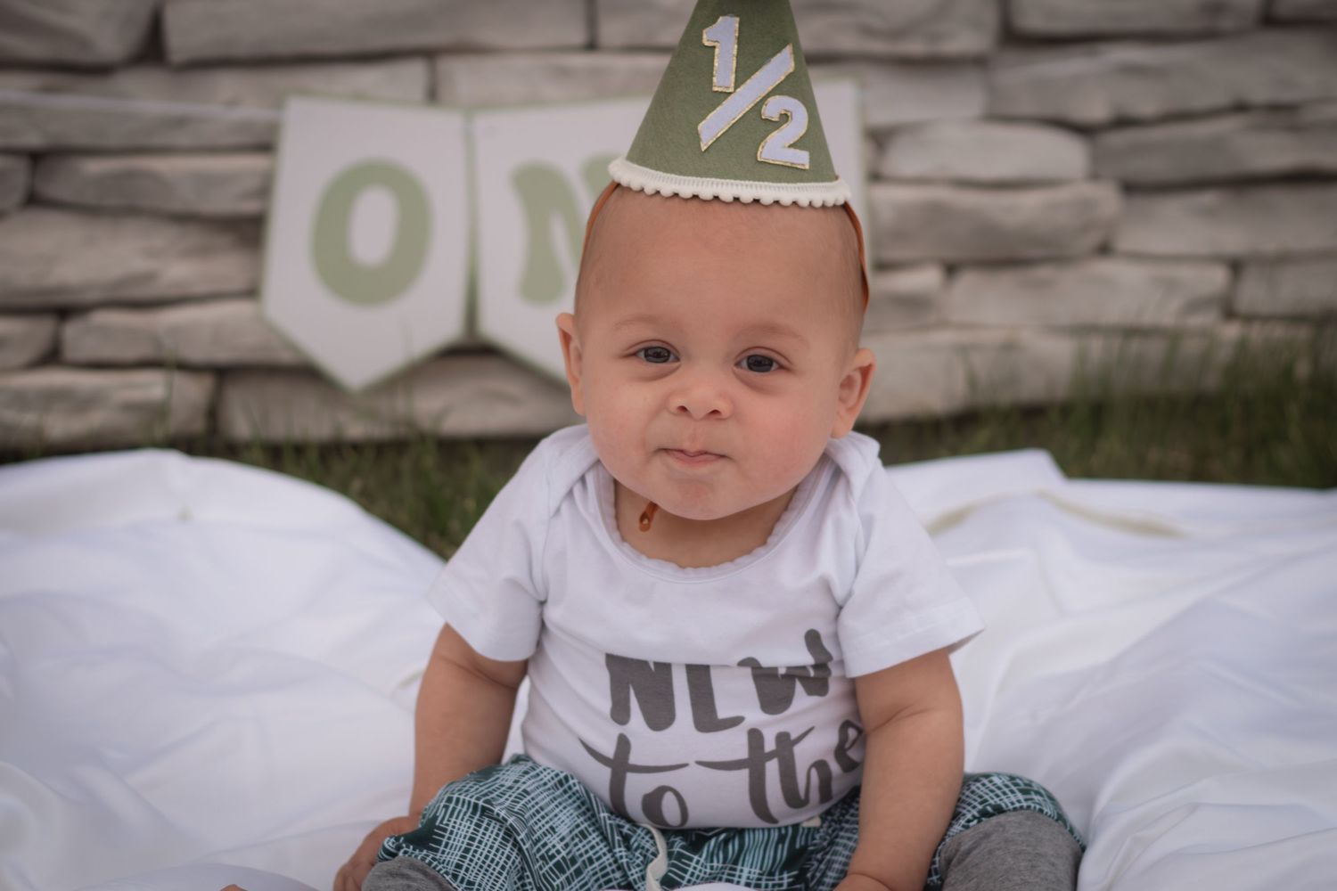 Baby in a half birthday hat and onesie sits on a white blanket outdoors celebrating their 6-month milestone.